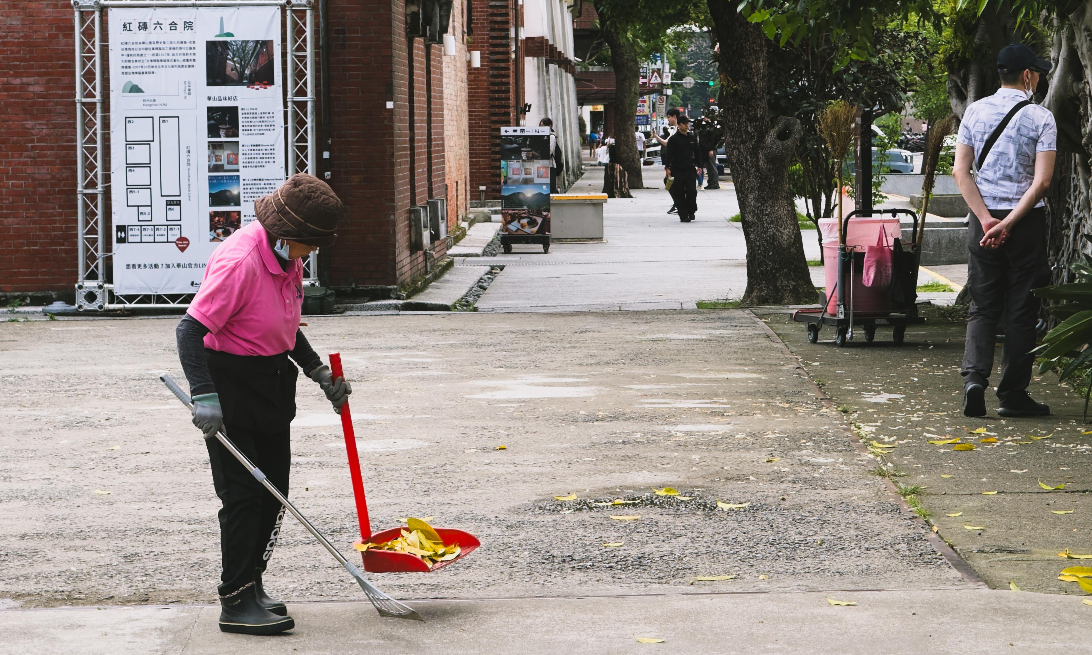A woman in pink janitorial uniform sweeps fallen leaves on sidewalk while a man in glasses and mask stands nearby with hands behind back.