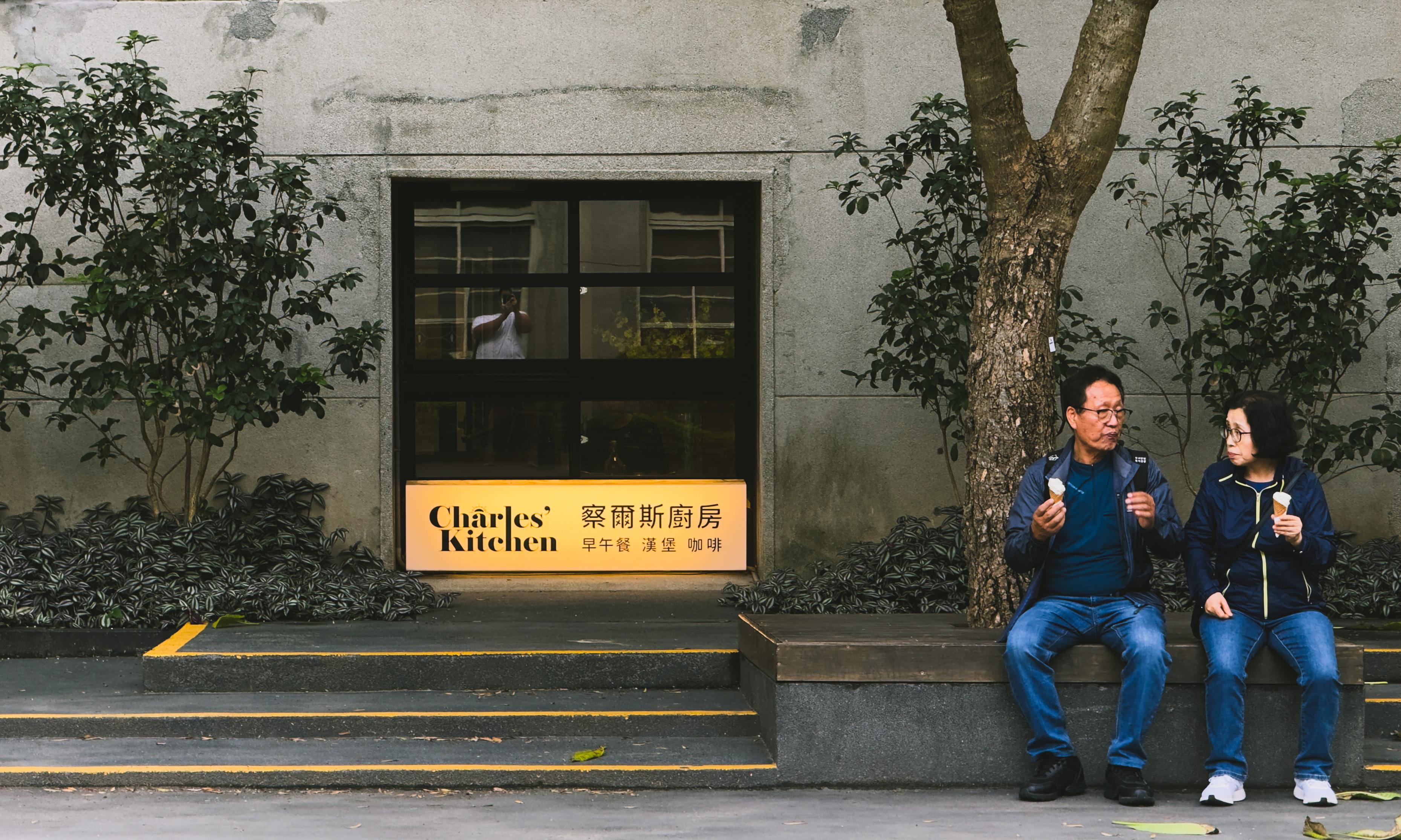 Two people sit on a bench eating ice cream outside a building with a sign that reads "Charles' Kitchen." One person is looking at the other while holding ice cream.