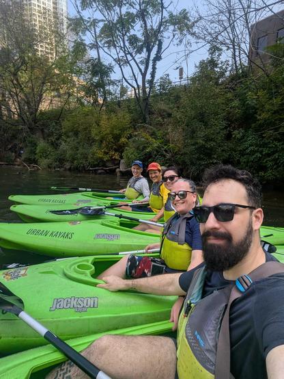 Five people smiling looking at the camera for a selfie in kayaks on the Chicago river with some greenery in the background. 
