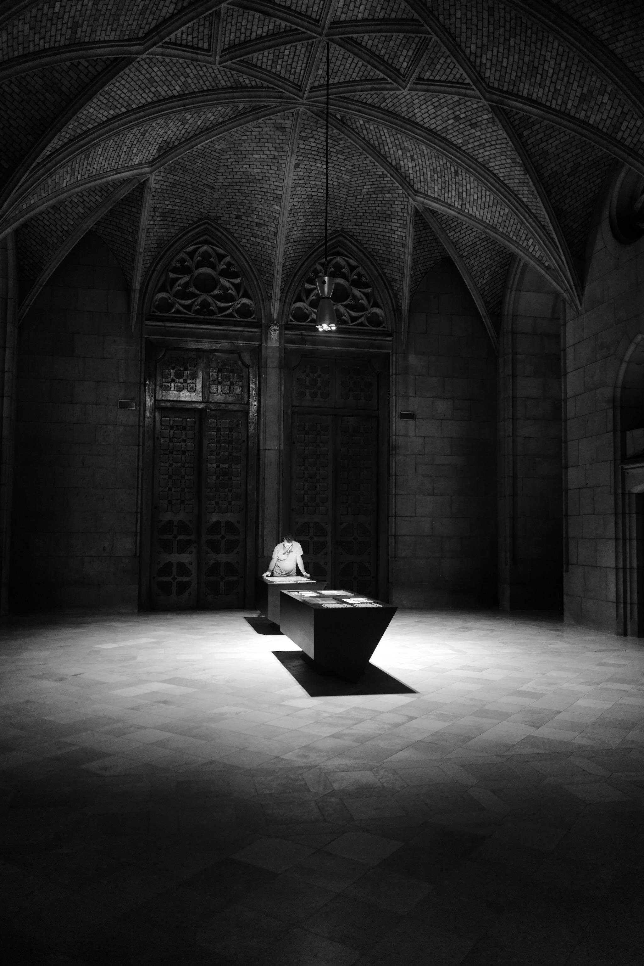 Picture of a person standing at a table. The table is located in a church (New Cathedral of Linz) under a cross vault. On the ceiling is a single spot, lighting the table and the person in circle. The person appears super bright in this dark room. The table throws a hard shadow in a rectangle shape on the floor.

Picture is black and white.
