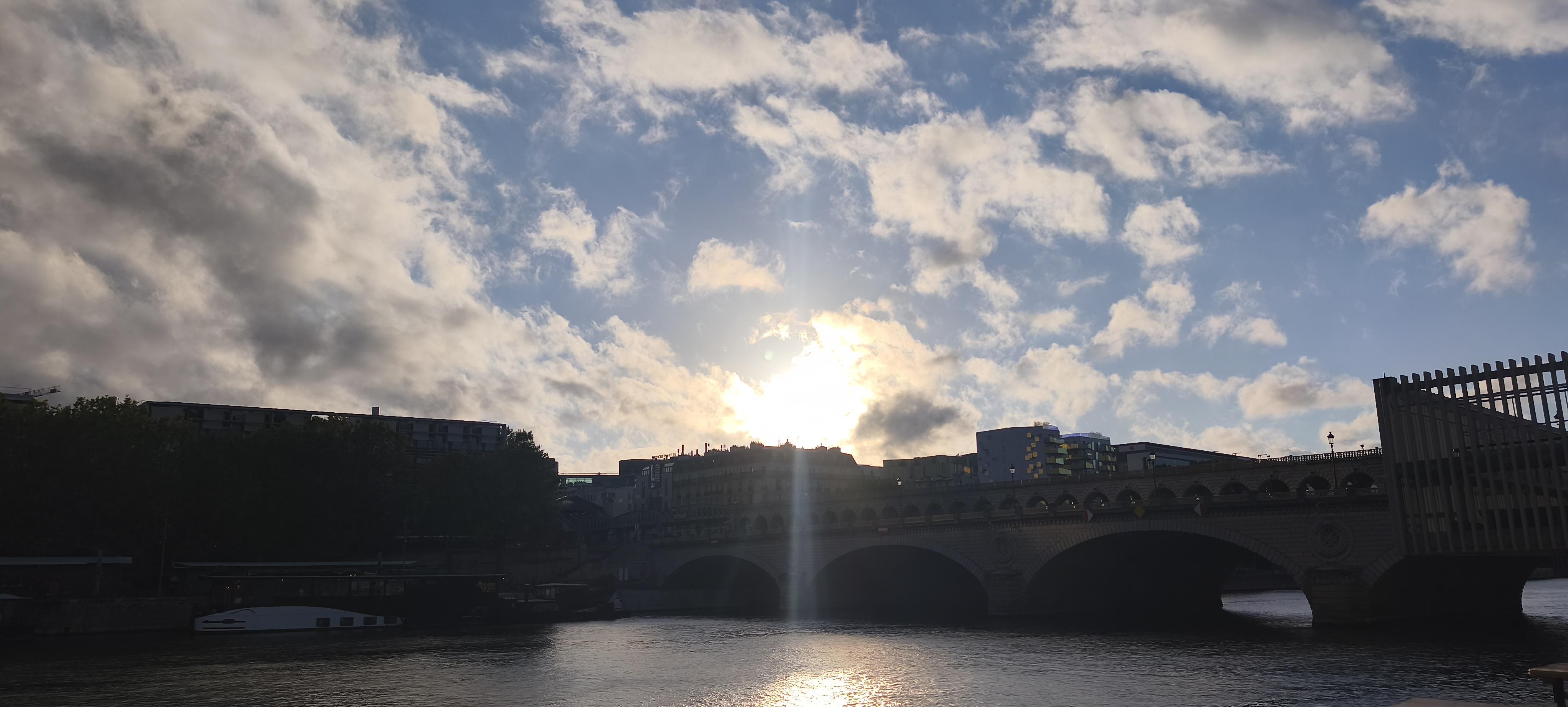 Vue sur la Seine au coucher du soleil, traversée par un pont aux grandes arches sombres. Le soleil perce entre les nuages et se reflète en une traînée lumineuse sur l’eau, tandis que le ciel bleu se teinte doucement d’or.