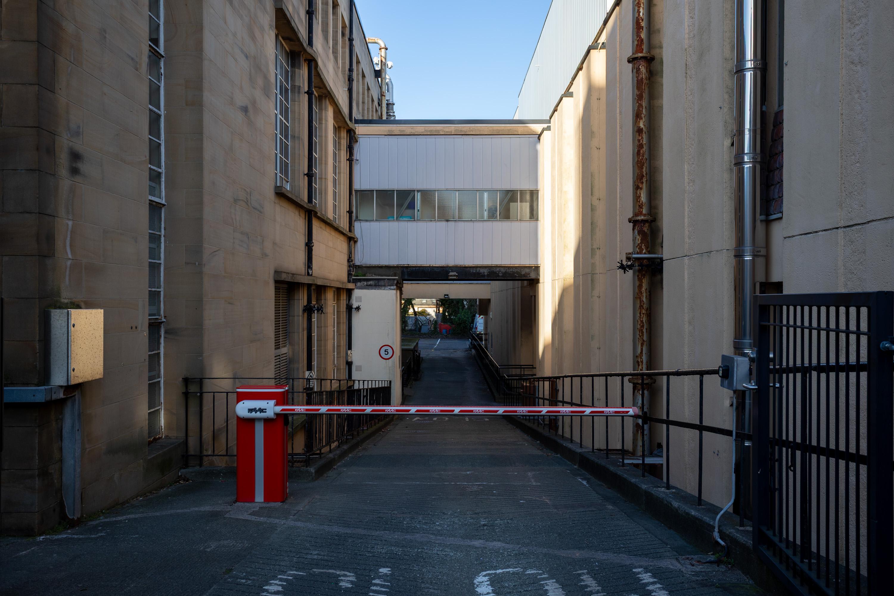 A narrow alleyway between two beige buildings with large windows, a red and white security barrier blocks entry. Overhead, a white enclosed walkway connects the buildings. The scene is lit by daylight and appears industrial.