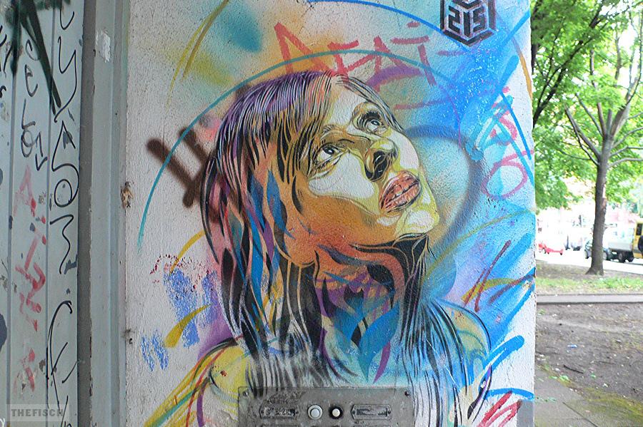 Ein Stencil einer jungen Frau. Es stammt von dem Pariser Künstler C215. Ein Porträt mit schulterlangen schwarzen Haaren mit blauen Sprähnen und verschiedenfarbigen Stencils auf hellem Hintergrund über einem Klingelschild. Der Blick der Frau ist nach oben gerichtet.
