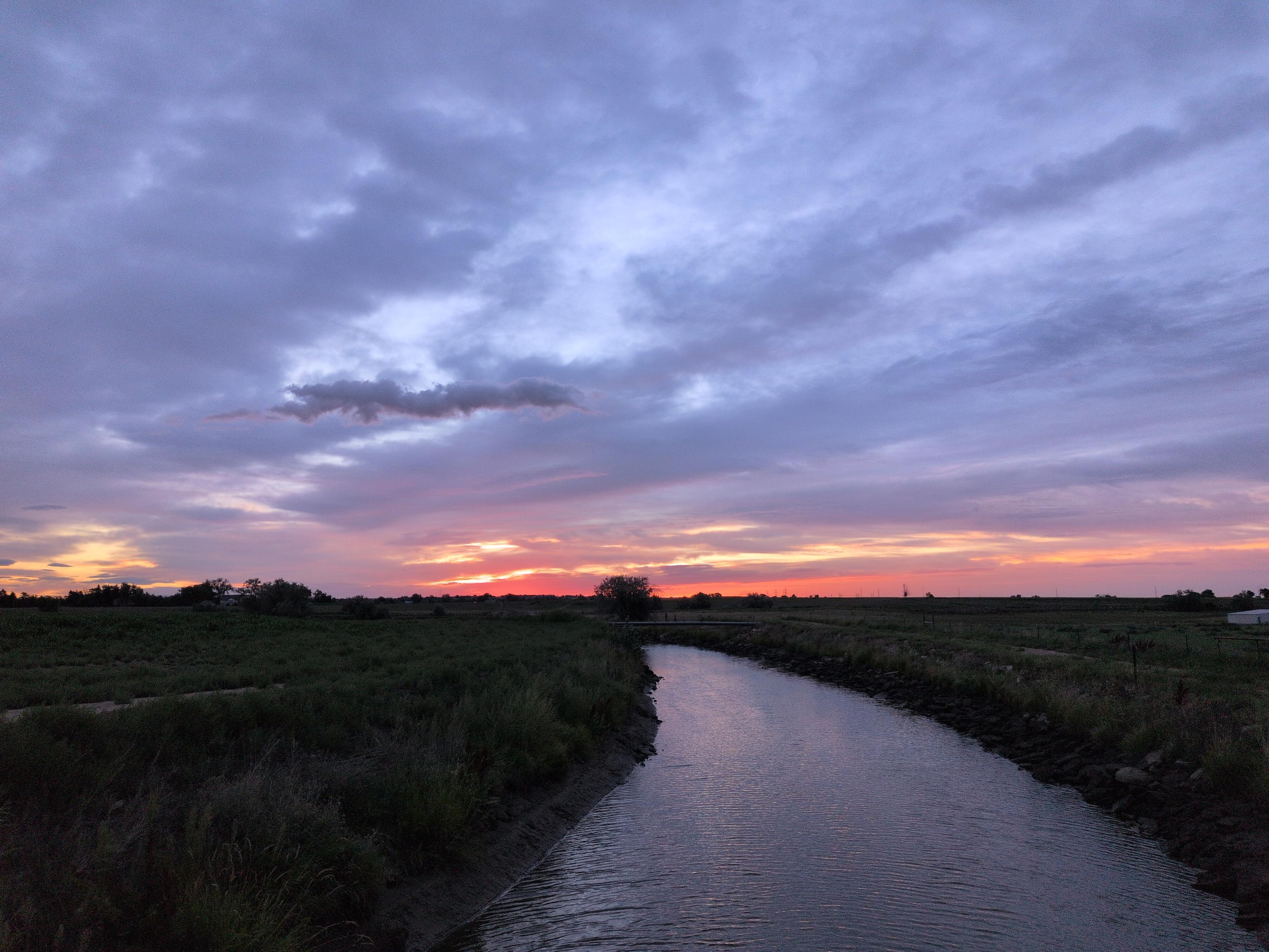 A canal runs out around a bend toward a sun which is beginning to redden, the water gently reflecting the faint blue-lavender of the skies above.
