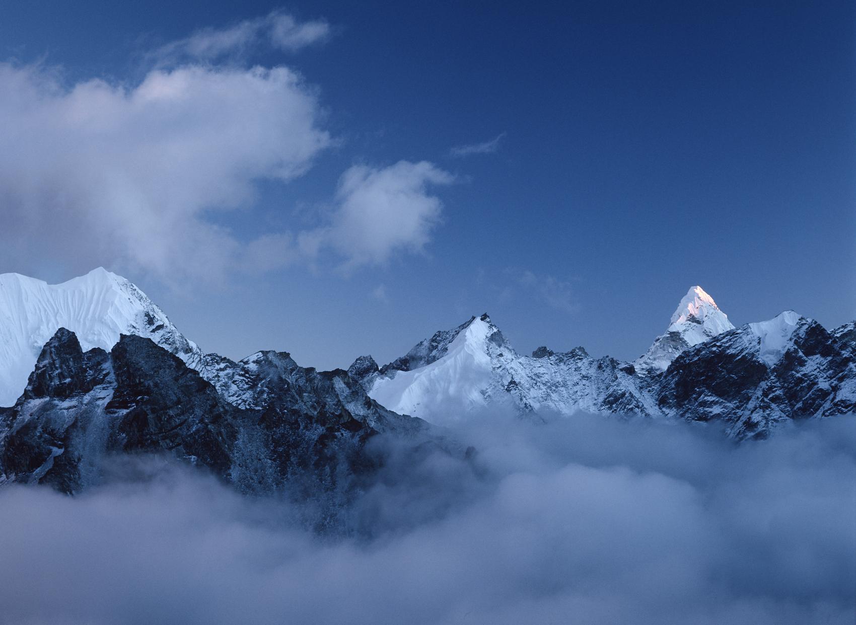 Snow-capped Himalaya mountains rise above a dense sea of white clouds. A sharp, prominent peak of Ama Dablam on the right is illuminated by sunlight against a deep blue sunset sky.