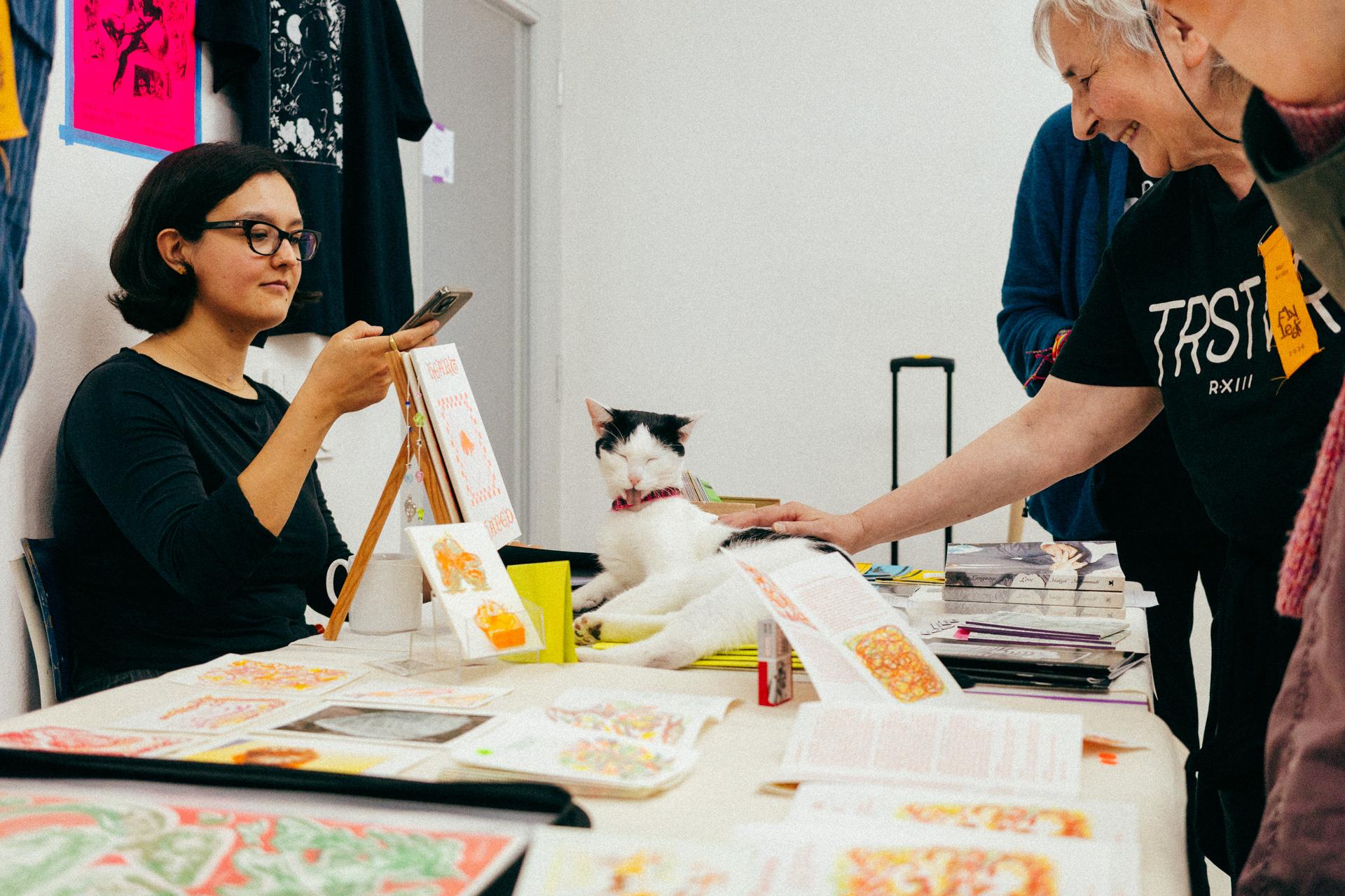 Fotografía de una linda gatita tumbada sobre un puesto de fanzines. A la izquierda de la imagen hay una persona haciéndole una foto, y a la derecha hay varias personas mirándola.

Photograph of a cute kitten lying on a fanzine stall. To the left of the image, a person is taking a photo of it, and to the right, several people are looking at it.