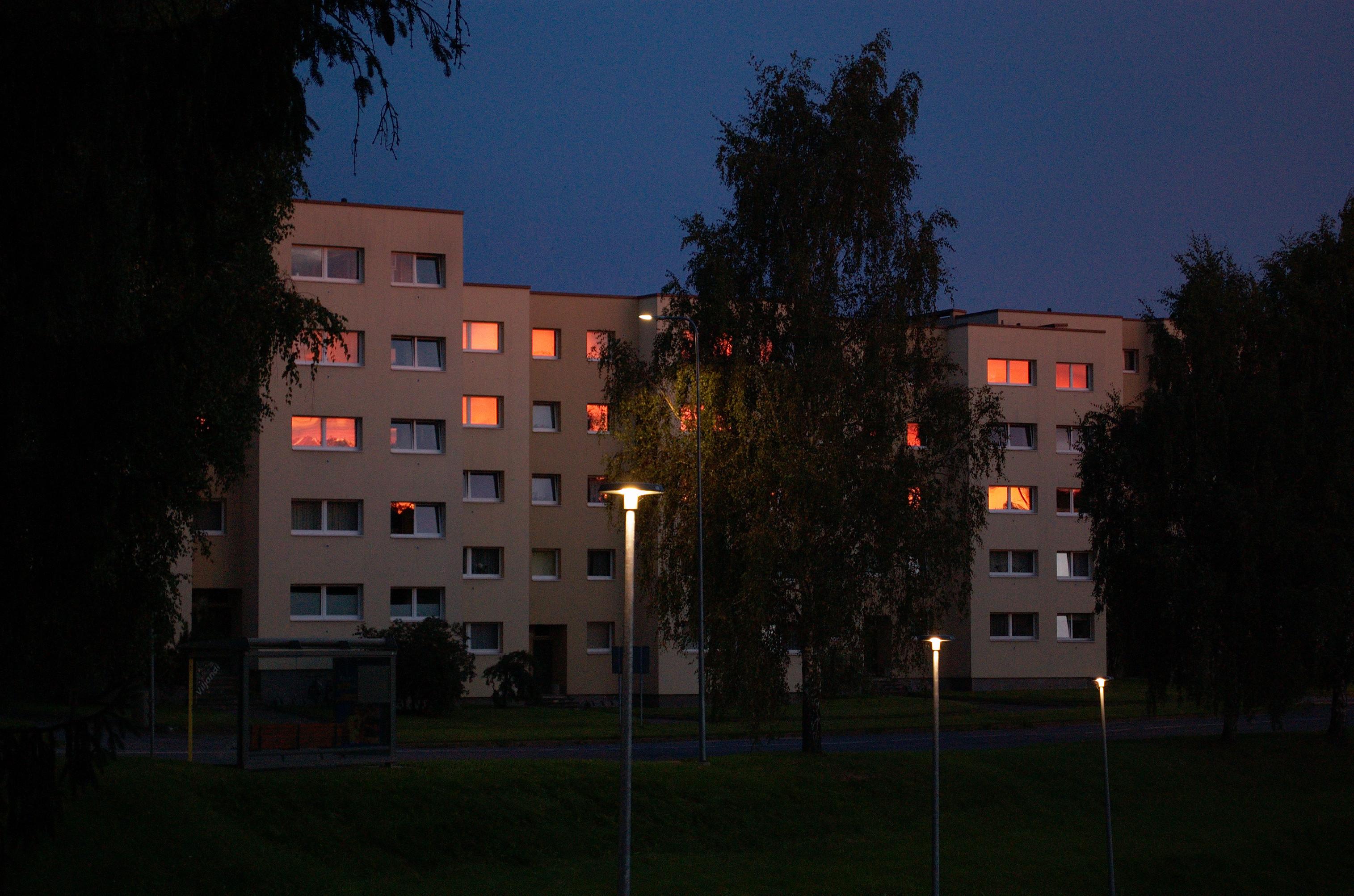 Photo of a six-storey building with a staggered facade, partially obscured by trees. It’s still quite dark and the sky in the background is deep blue, but the rising sun casts a pink glow on the building and many of the upper windows gleam orange and red.