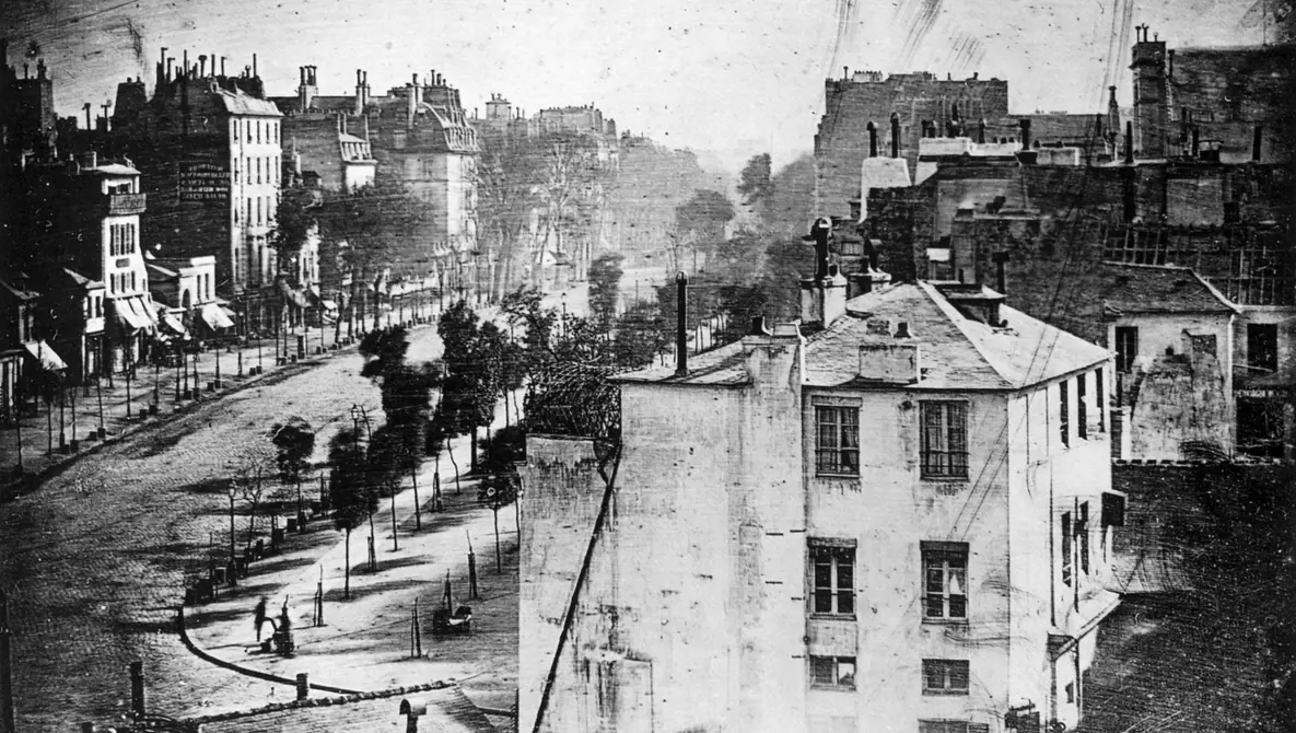Fotografia in bianco e nero di un ampio viale urbano alberato, con binari e pali sul lato della strada, circondato da edifici ottocenteschi alti e densamente costruiti. In lontananza il viale prosegue tra gli edifici e la vegetazione; in primo piano sono visibili tetti, camini e facciate di abitazioni. Una sola persona è visibile, apparentemente mentra si lucidare le scarpe.