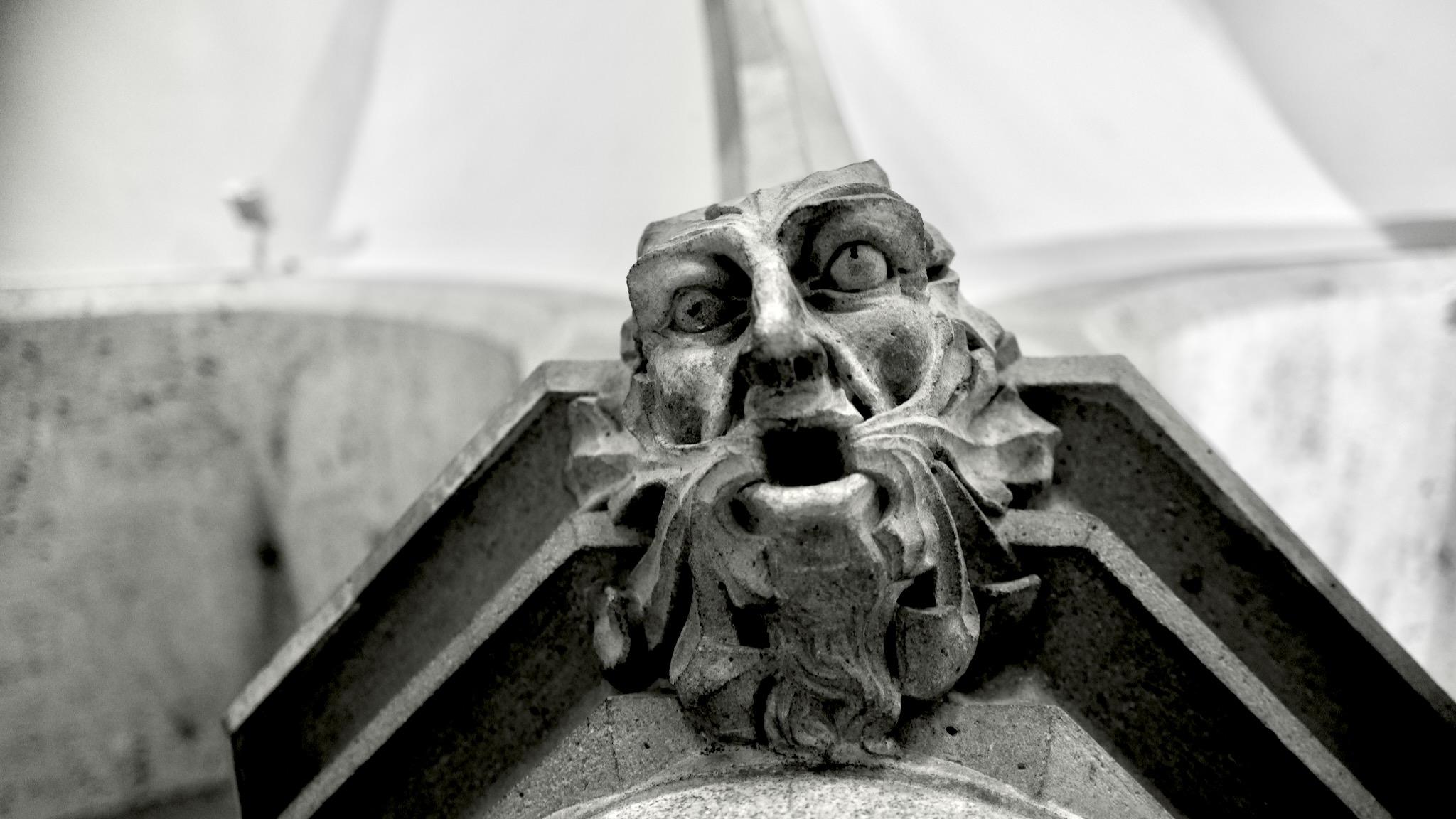 Picture of a stone face grimace on a pillow in the crypt of the New Cathedral in Linz. It is formed as a bearded man, mouth opened, looking down on the spectator. The beard hair is whrily and layered in waves around the head. In the background are lines from the pillar. Photo is black and white.