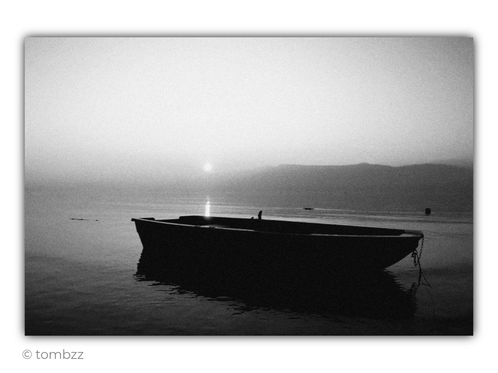 Black-and-white analog photograph of a small boat floating on calm water at sunset. Its dark, almost featureless silhouette occupies the lower part of the frame, standing out strongly against the lighter surroundings. A chain hangs from the bow, while the boat’s reflection softly dissolves across the gently rippled surface.
In the distance, a dark line of low hills stretches along the horizon, above which the sky fades through subtle shades of gray. A small, dimmed sun hangs low near the horizon, casting a narrow vertical trail of light across the water and drawing the eye deeper into the scene. The analog grain and soft tonal transitions give the image a melancholic, almost timeless quality. The composition feels quiet and restrained, built around stillness, space, and the contrast between the heavy darkness of the boat and the luminous water and sky. The whole scene feels like a moment suspended just before dusk.
