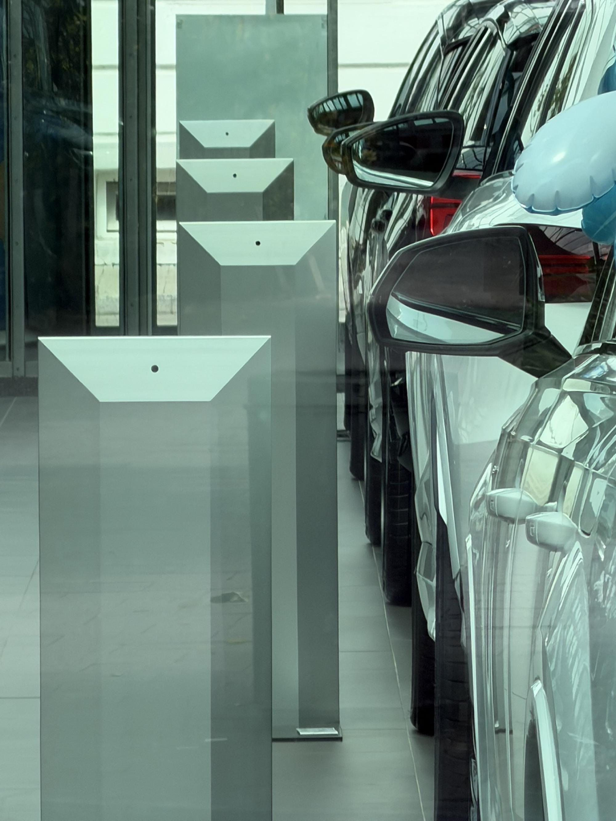 A row of cars stands closely aligned inside a showroom. Their side mirrors repeat toward the back of the image, while a parallel row of pale geometric display stands recedes on the left.