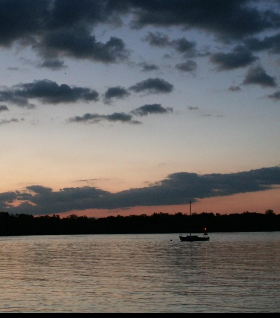 Dark lake with a boat in the distance, tree lined coast, and a pink sky with clouds, below sunsetting skies