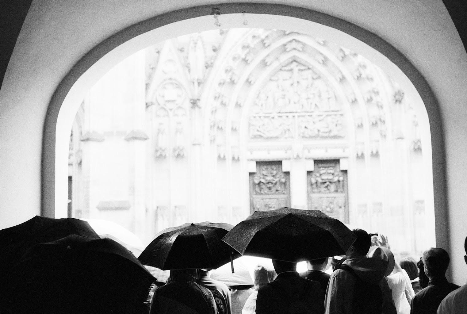 A group of people holding black umbrellas stands in an archway, facing a detailed gothic-style doorway. The scene is monochromatic, conveying a rainy atmosphere.
