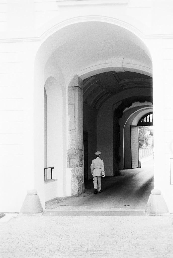 A solitary soldier in uniform walks through a large archway, leading into a corridor with muted light. The image is in black and white, highlighting the architectural details of the passage.