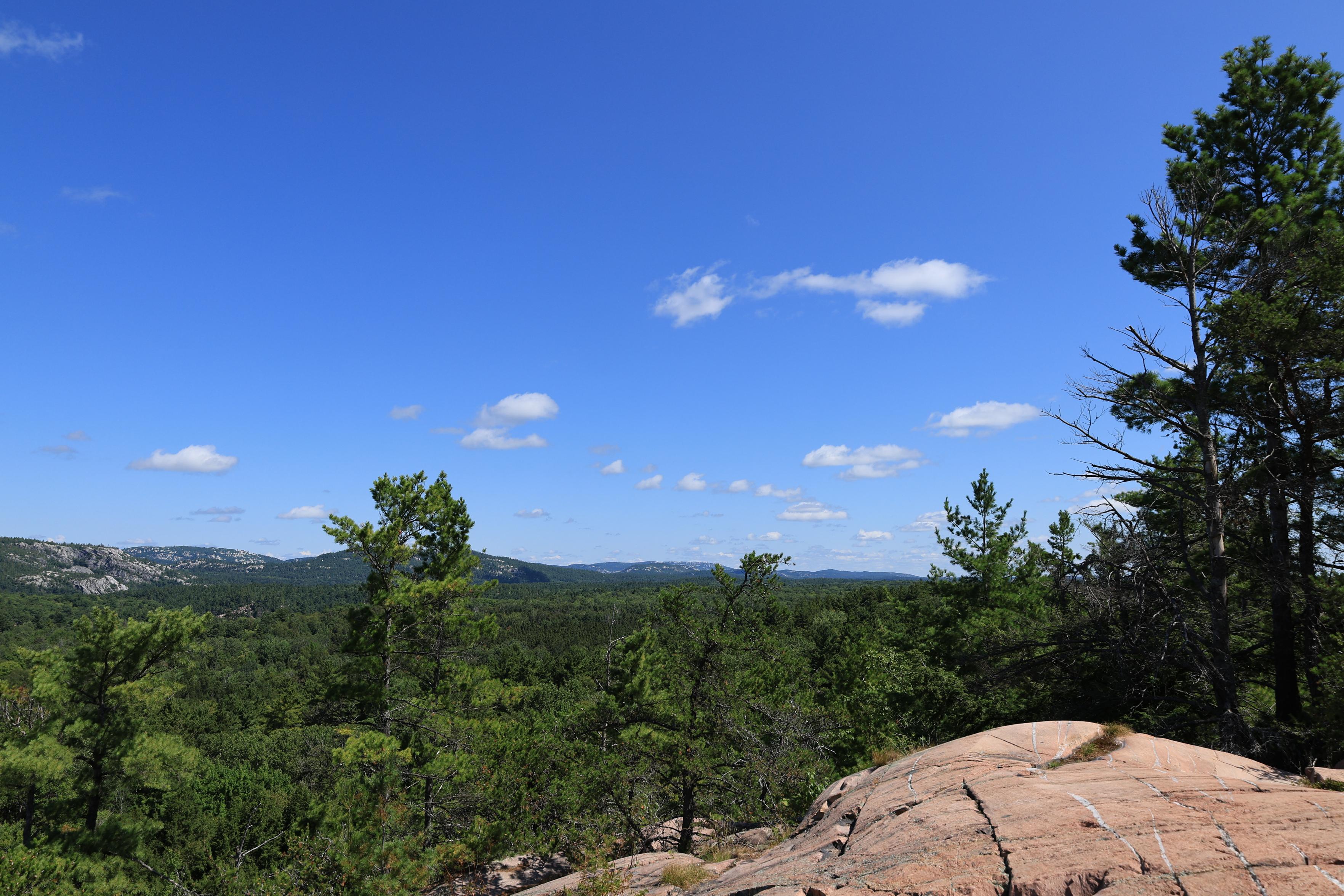 This is a landscape photo taken during the summer from a lookout at the top of a hiking trail. An outcropping of granite rock is in the foreground with a forest extending in the distance. A lower level mountain range can next be seen in the distance. The sky above is a vivid blue with only a few white clouds in view.