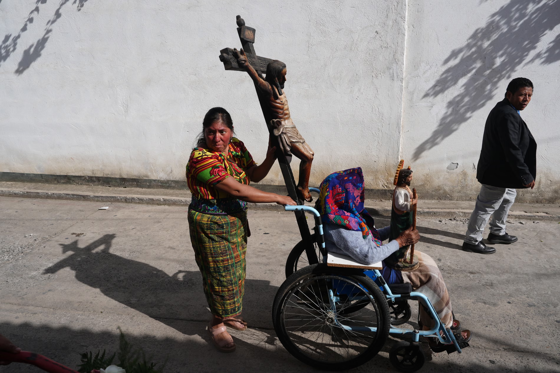 A woman in vibrant traditional Indigenous clothing pushes an elderly person sitting in a light blue wheelchair along a paved street past a plain white wall. A large wooden crucifix with a carved figure of Jesus is attached upright to the back of the wheelchair, and the person in the chair holds a small religious statue of a saint.