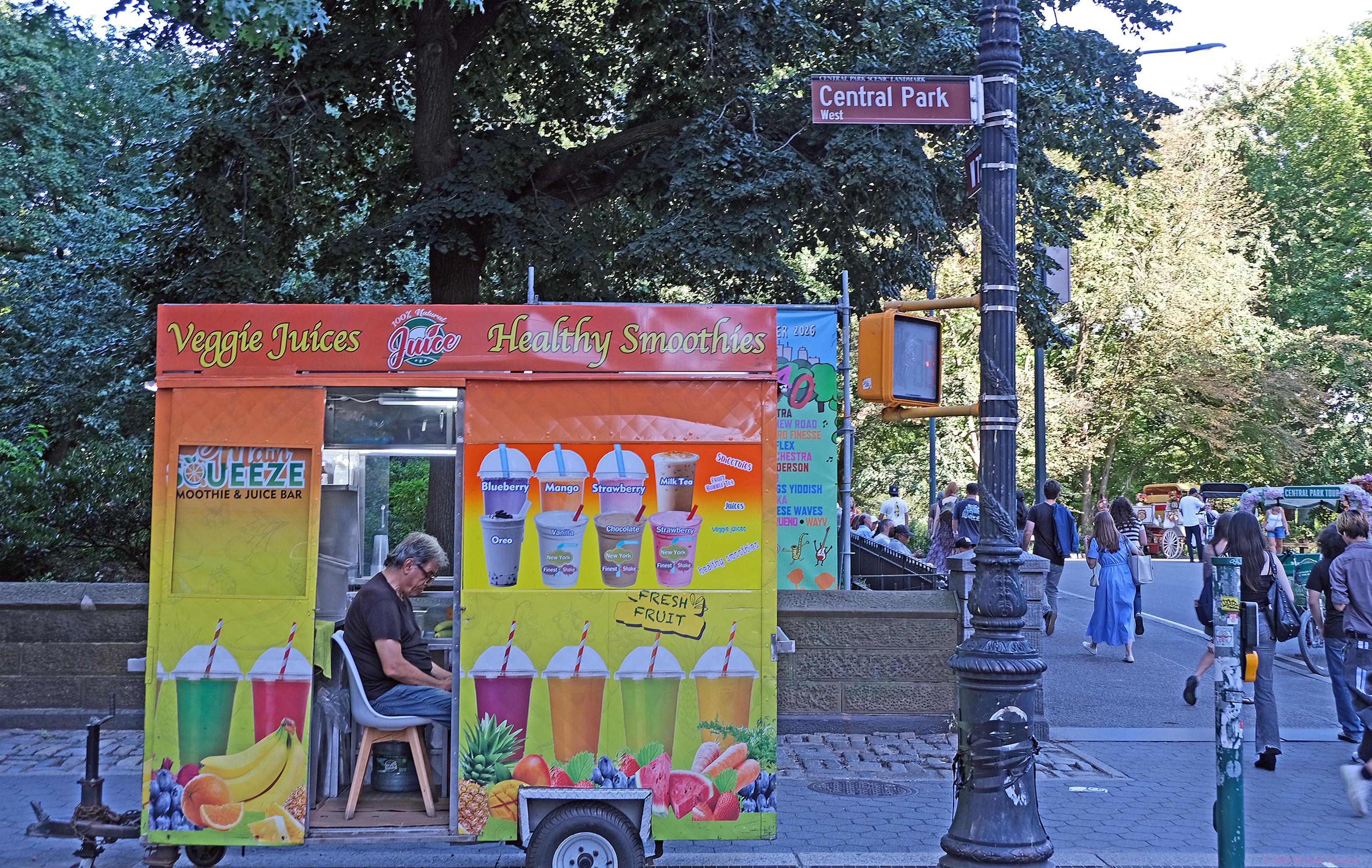 Photo by Fenichel:
The view from the edge of the entrance to Central Park on Central Park West (the "Women's Gate"). On the left half is a colorful food truck/trailer offering smoothies and Veggie Juices. In the middle of the cart's wall, with images of all the drinks is a doorway, in which the vendor is framed, sitting on a chair.

The right half features the sign post in front of the gate (leading to "Imagine" and "Strawberry Fields") and people are walking into the park, with a "Central Park Tour" bicycle taxi and a horse-drawn carriage nearby.