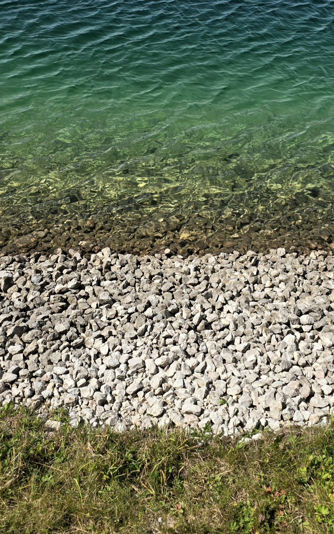 Tag. Sonnig. Künstlicher Bergseerand. Erst Gras, dann helle Schottersteine, dann Wasser hellgrün bis dunkelgrün.