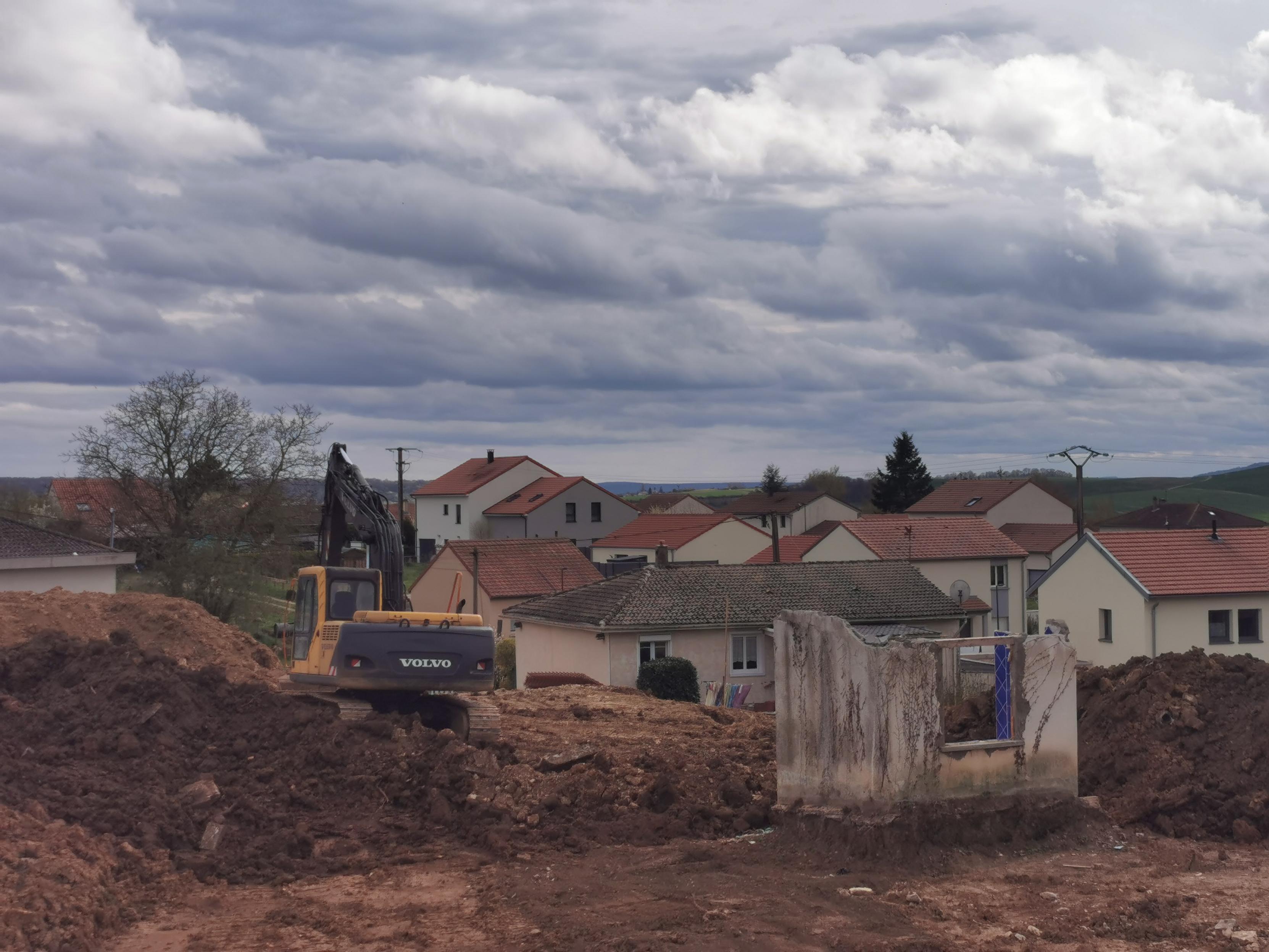 Chantier, pelleteuse, terre retournée, ruine, dans un village sous un ciel tourmenté.