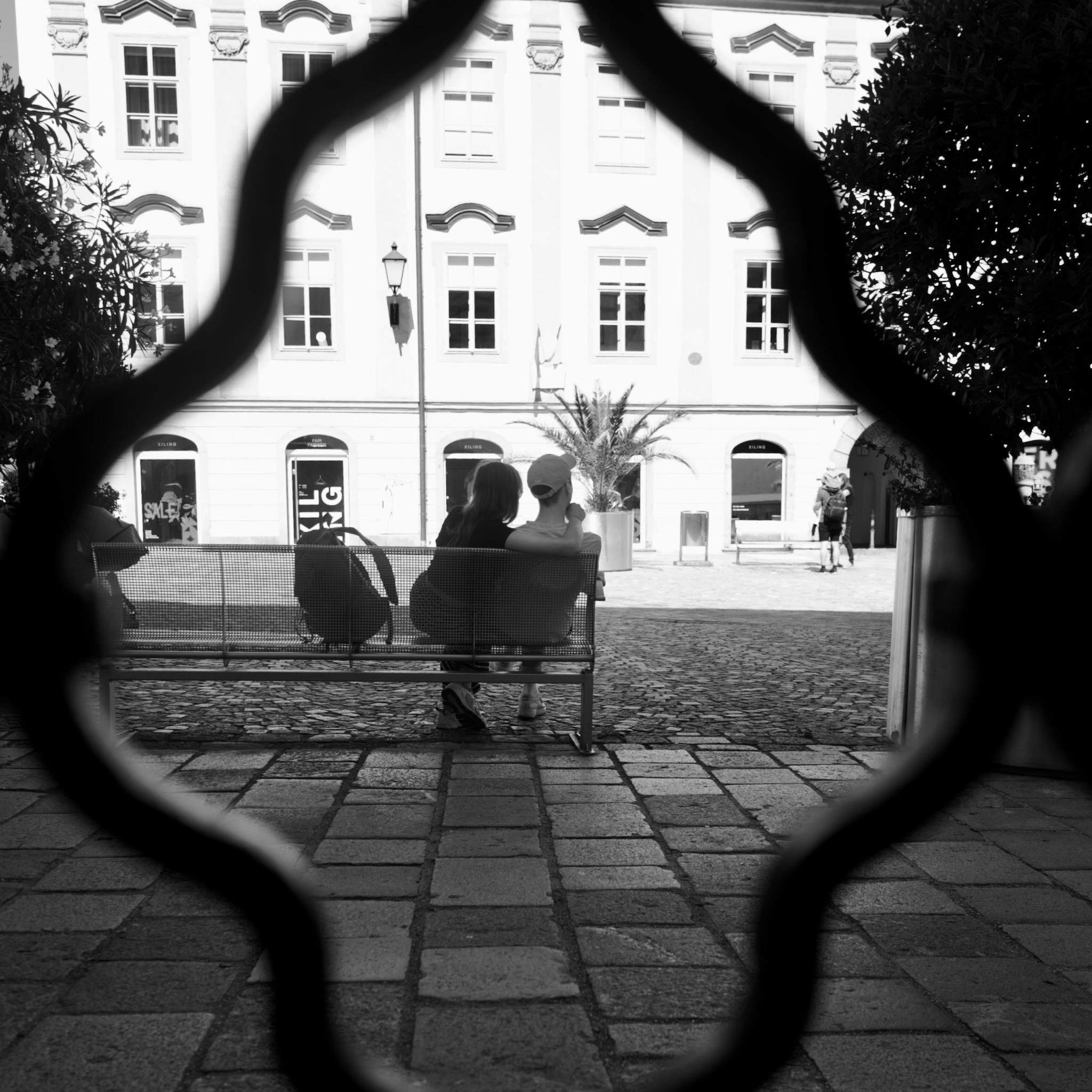 Picture of a young couple sitting on a bench in Linz, Austria. She is sitting on the left hand, leaning to him. He is wearing a white shirt and a white cap, she is dressed in dark colors. Next to her is a backpack on the bench.
In the background is a bright classical facade of a building. In the foreground are some plaster stones. 
Left and right are big plants framing the picture. In the very foreground is a blurred metal grid giving a diamond shaped curved frame.