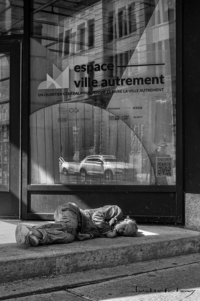 A person sleeping on the ground in front of a building . A sign in the window says : "Espace Ville Autremebt" or"A Different City Space"