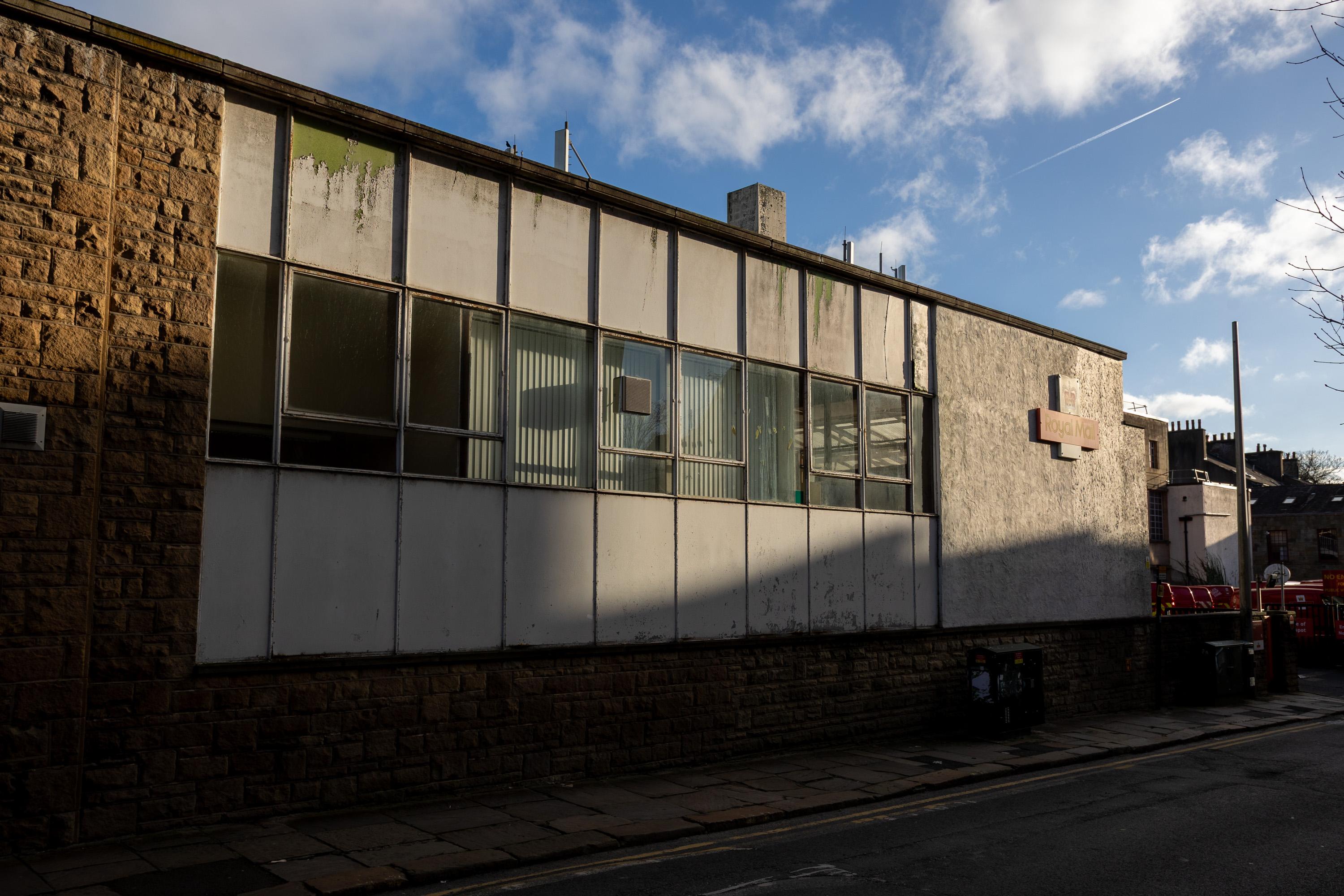 A sunlit, old building with large, grimy windows, white panels, and a stone base lines a quiet street with bins and shadows. The sky is blue with clouds and a visible plane vapour trail.