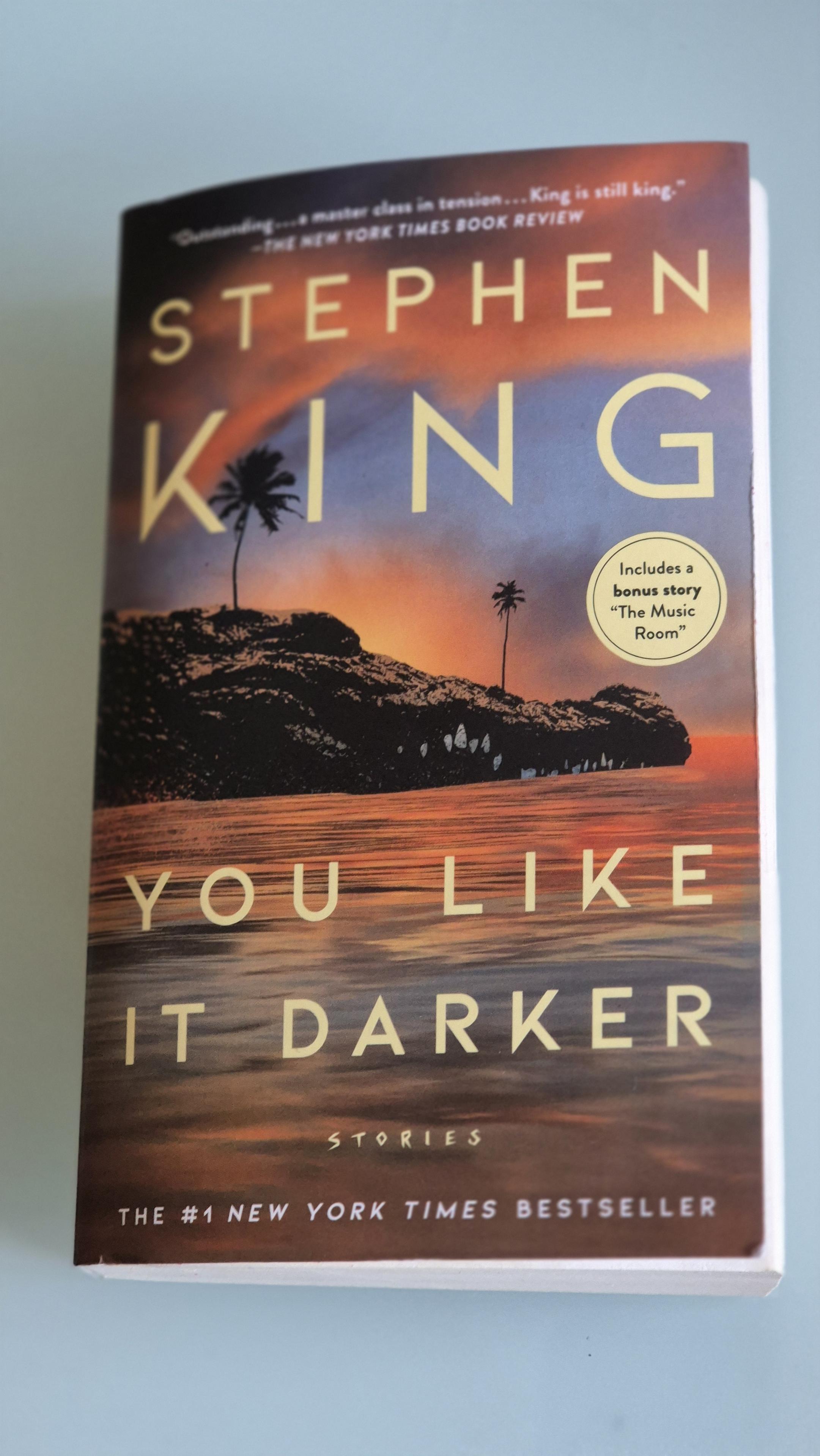 Das Foto zeigt das Cover von Stephen King's Storysammlung "You Like It Darker". Es weist eine felsige Insel, auf der zwei Palmen stehen, auf. Das Wasser spiegelt einen grandiosen Sonnenuntergang. /
The photo shows the cover of Stephen King’s short story collection *You Like It Darker*. It depicts a rocky island with two palm trees on it. The water reflects a magnificent sunset.