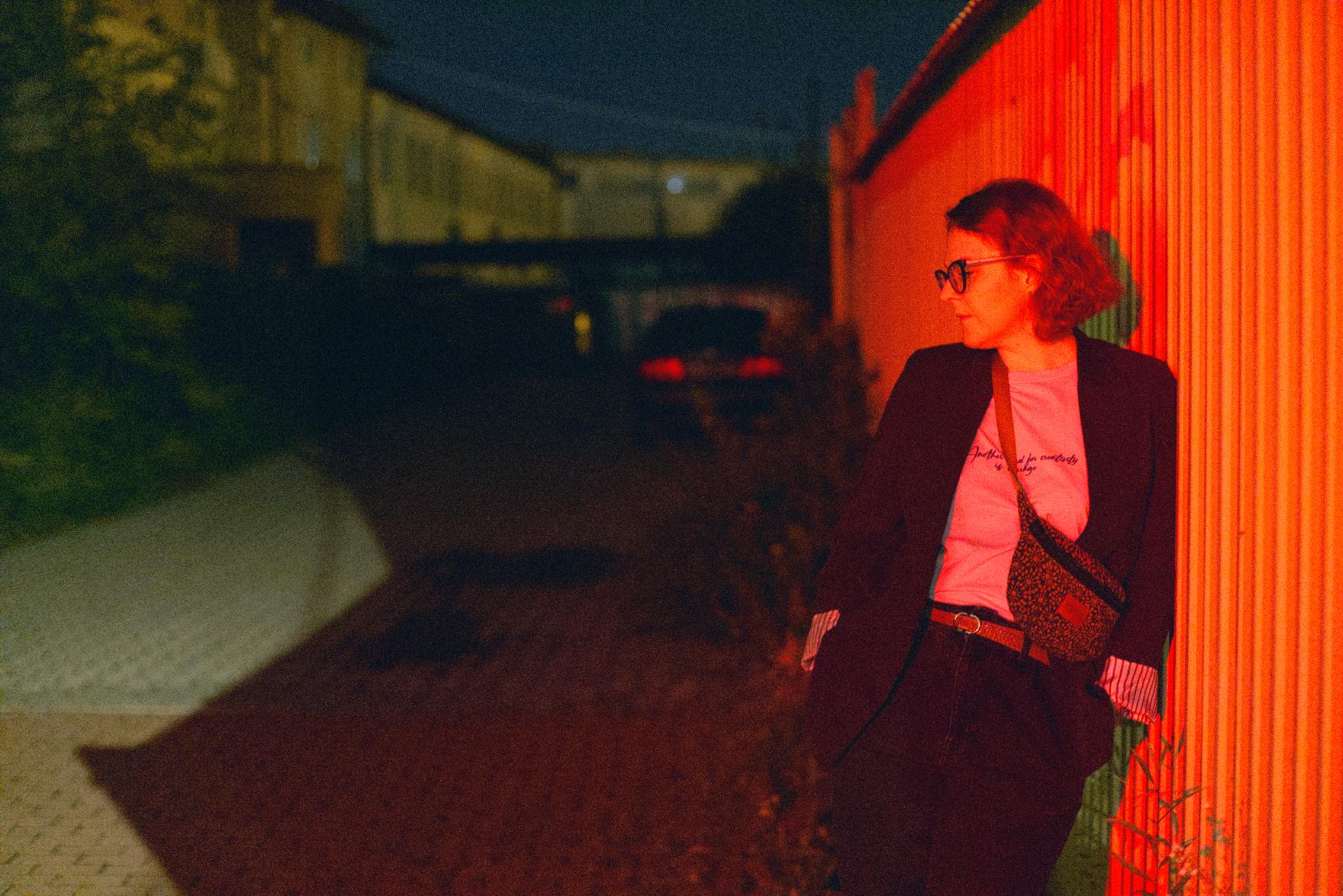 To nocna fotografia, na której po prawej stronie zdjęcia kobieta opiera się o metalowy płot, oświetlony czerwonym światłem. Patrzy w lewo. Jej wzrok skierowany jest na stojący w cieniu, przed jakąś bramą, samochód. Dalej, na tle granatowego nieba stoją żółte domy, a po lewej stronie zarośla.