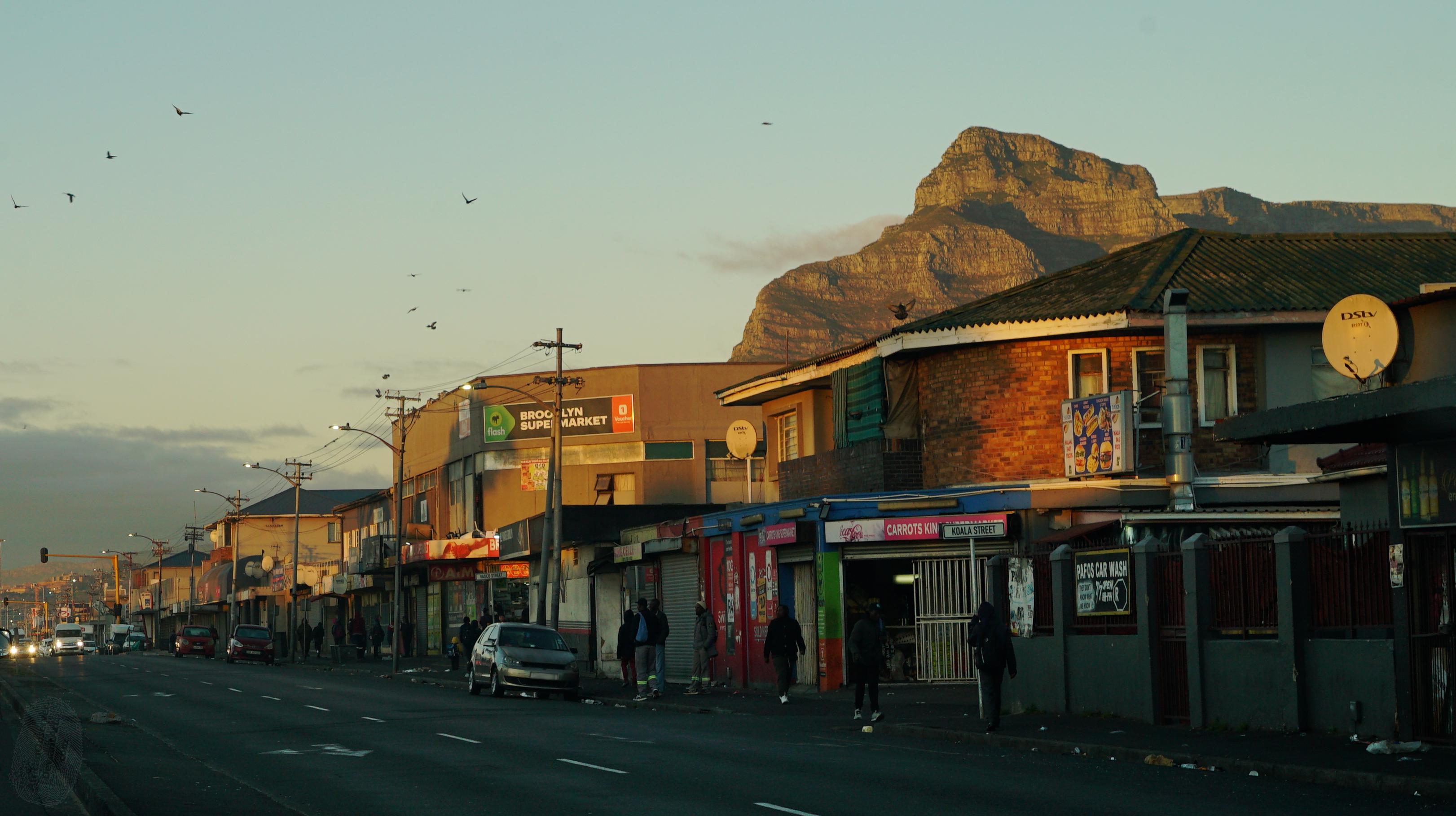 A city street in warm sunrise light, warm coloured buildings down the right of an otherwise empty road. Vehicles with lights on can be seen in the distance. Off to the right is a mountain