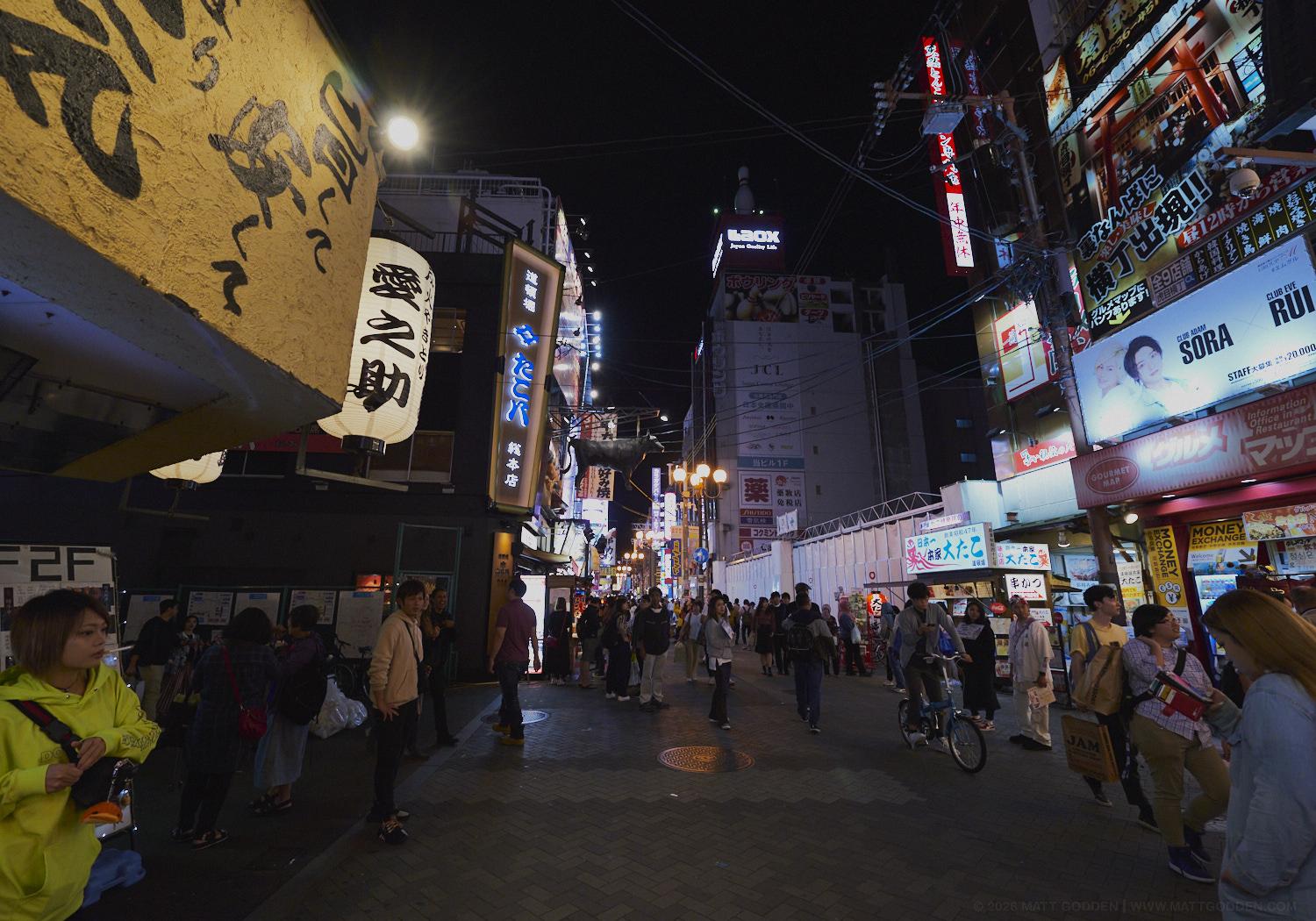 A street scene in crowded Dotonburi Osaka. Lit signs and billboards and cables stringing across the space, people on bikes, looking at phones, holding signs for restaurants, and in the middle distance, across a clearing in the crowd, is a man standing dead-still, staring straight at me.