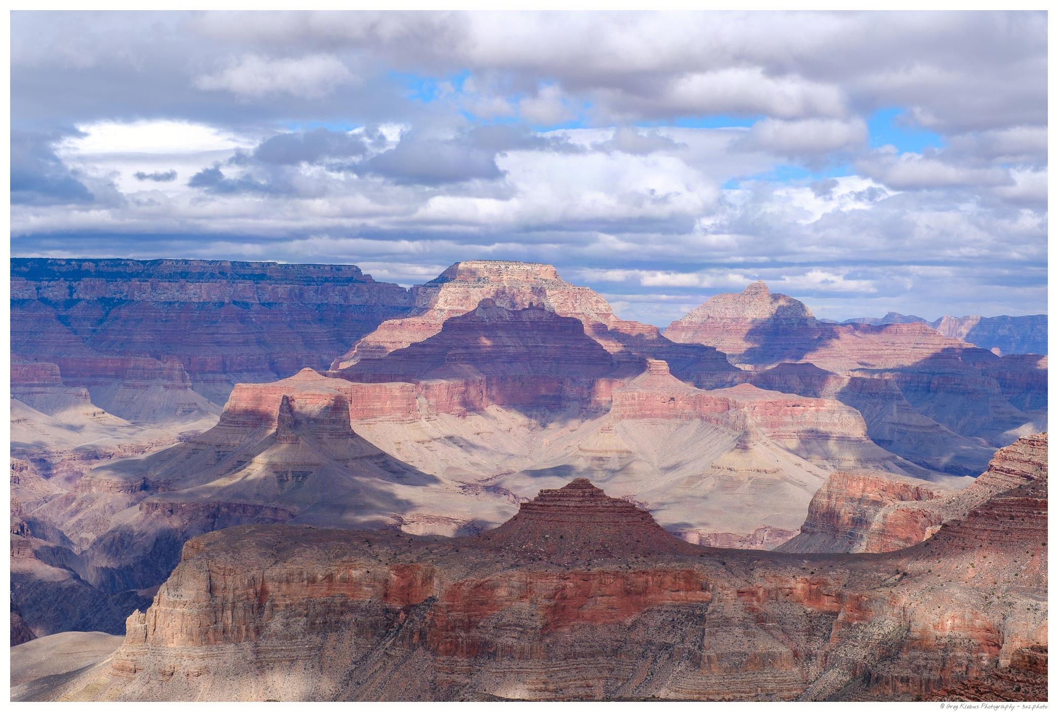 A wide view of the Grand Canyon, showing layered red-rock cliffs and mesas beneath a dramatic sky filled with clouds.