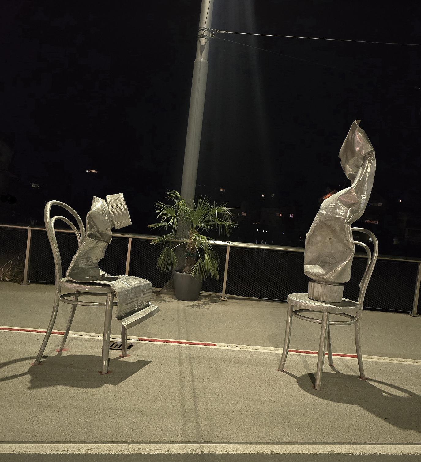 Nacht. Unter einer hellen Straßenlampe. 2 Kunstskulpturen. Geknautschte Tuben sitzen gegenüber zugewandt auf Caféstühlen.