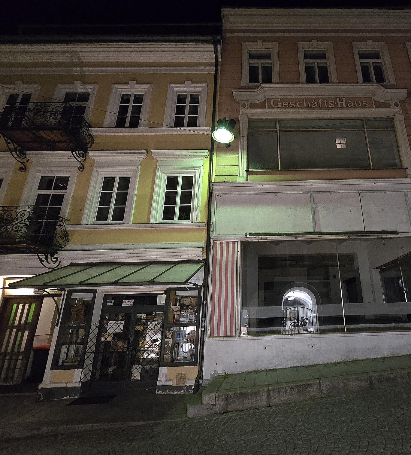 Nacht. Farbig. Ein leeres Geschäftshaus. Daneben ein alter Buchladen. In der Mitte eine helle Straßenlampe. In den Scheiben spiegelt sich die gegenüberliegende Straßenseite.