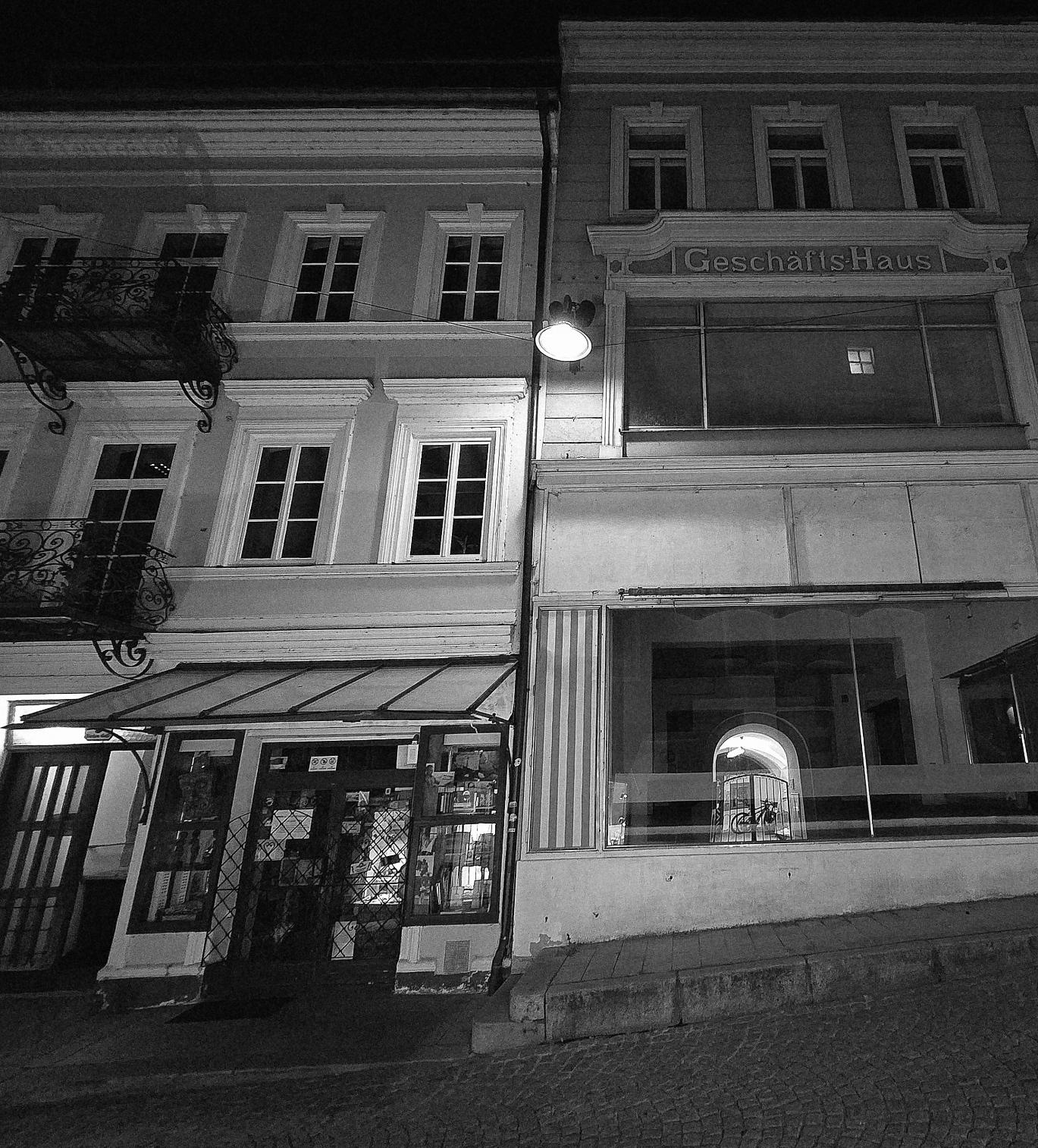 Nacht. Schwarzweiß. Ein leeres Geschäftshaus. Daneben ein alter Buchladen. In der Mitte eine helle Straßenlampe. In den Scheiben spiegelt sich die gegenüberliegende Straßenseite.
