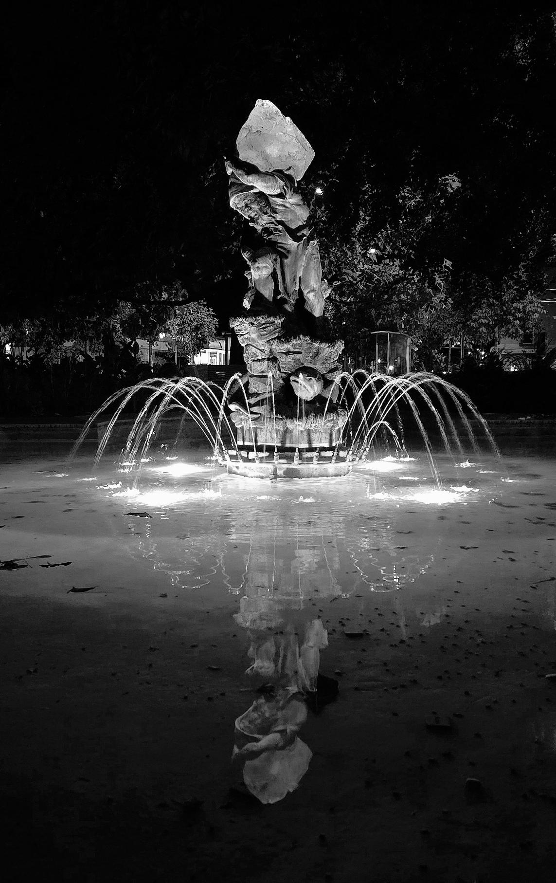 Nacht. Schwarzweiß. Springbrunnen. In der Mitte eine Skulptur. Ein Gnom, der einen sehr großen Bergkristall trägt. Im Wasser spiegelt sich der Gnom.