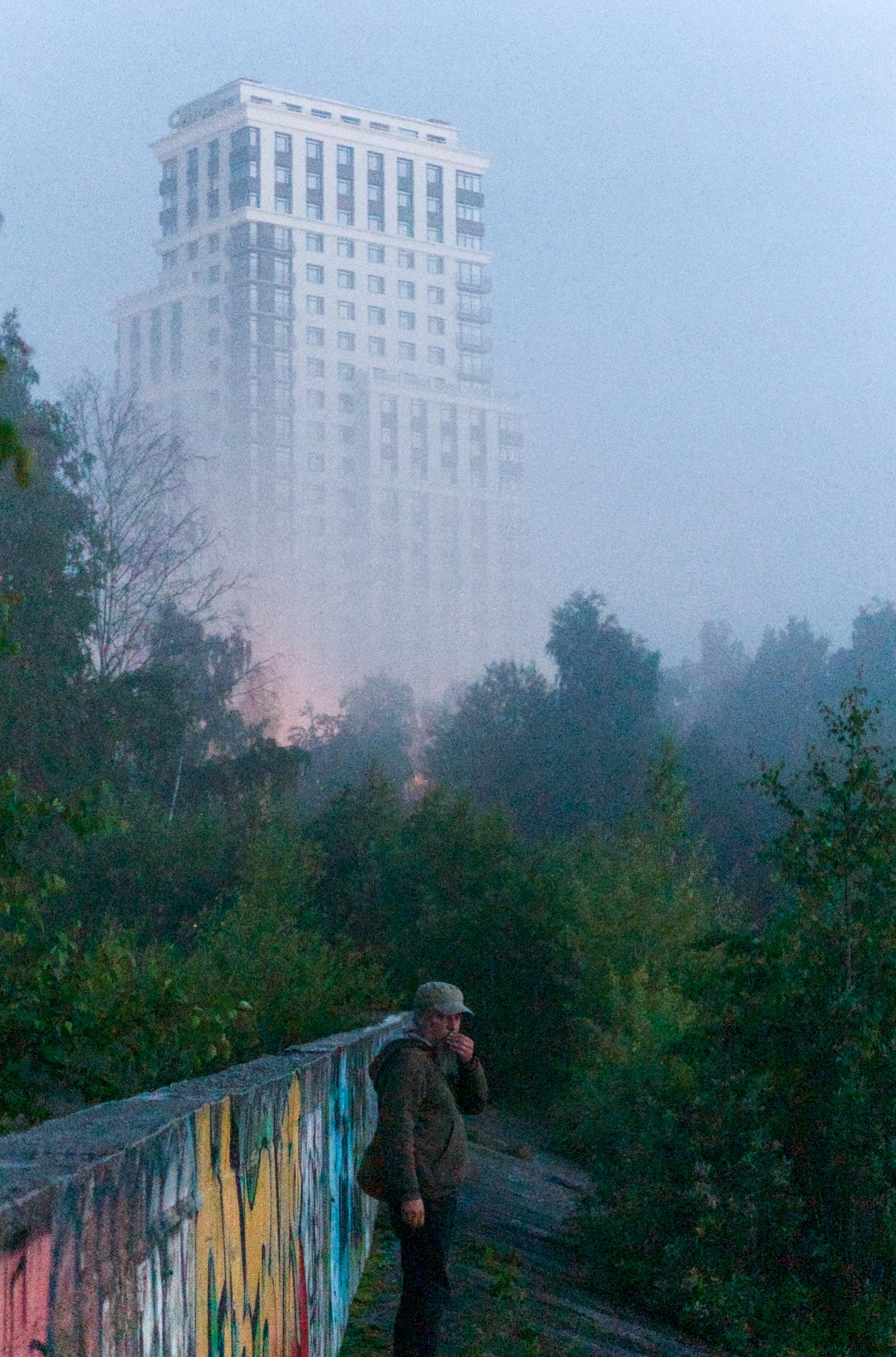 This color photograph captures the atmosphere of thick fog in the urban jungle. In the center of the frame stands a solitary man wearing a cap and a dark jacket, standing next to a concrete wall covered in colorful graffiti. He is smoking and looking intently toward the camera, creating a sense of alertness. Behind him, dissolving into a dense milky haze, is a tall modern residential building, which appears as a ghostly mirage due to the fog. The space between them is filled with dense, wild overgrowth of bushes. The image conveys a sense of anxiety, mystery, and a post-apocalyptic atmosphere, where a familiar city transforms into a mysterious "Zone."