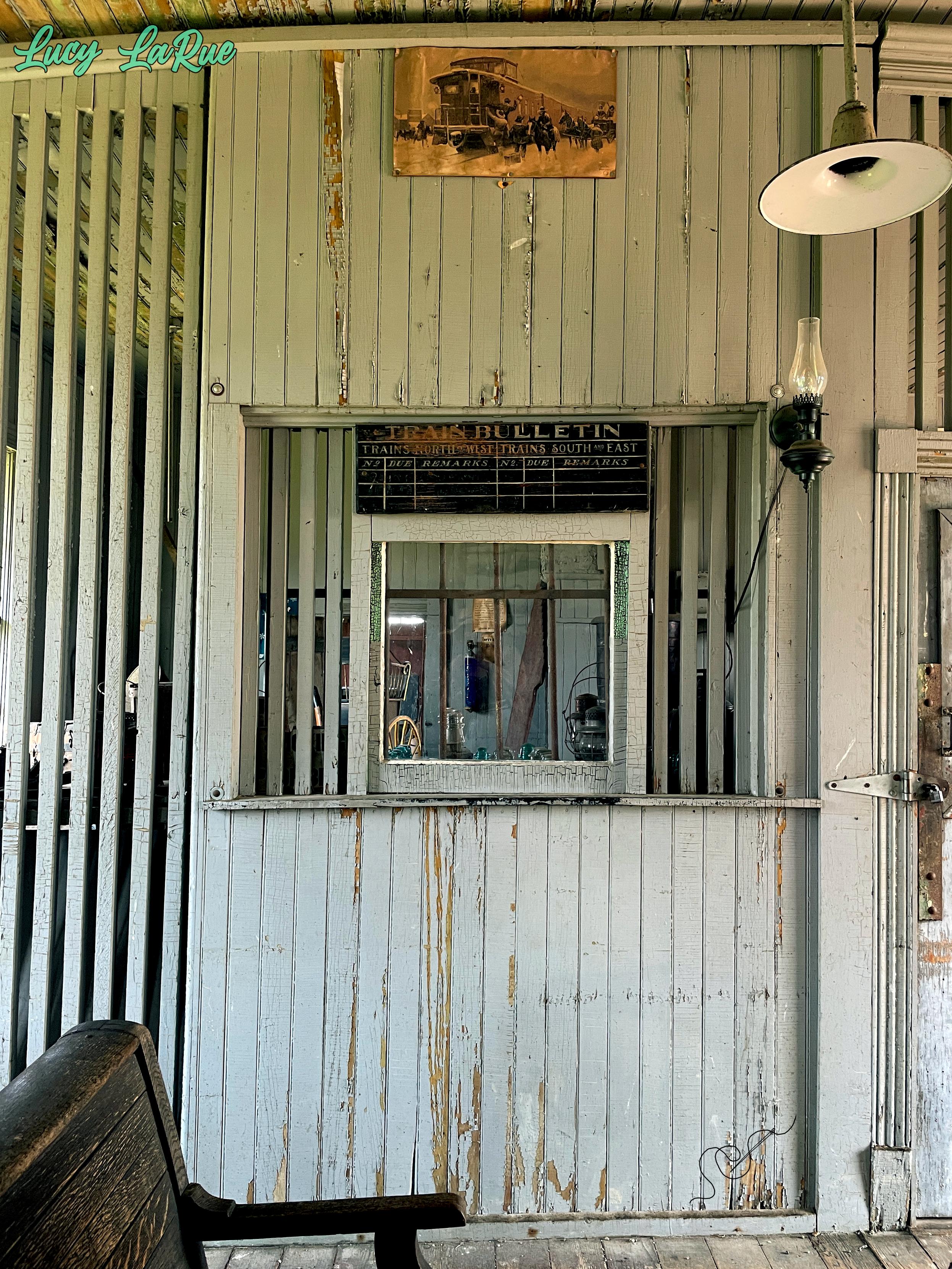 A vintage train depot ticket window, dusty teal-grey wooden slats framing the opening back into the retro ticket office, the paint chipping significantly with age. A dark wooden seating bench is in the foreground, and a sepia photo of a train arriving at the station is posted near the ceiling.
