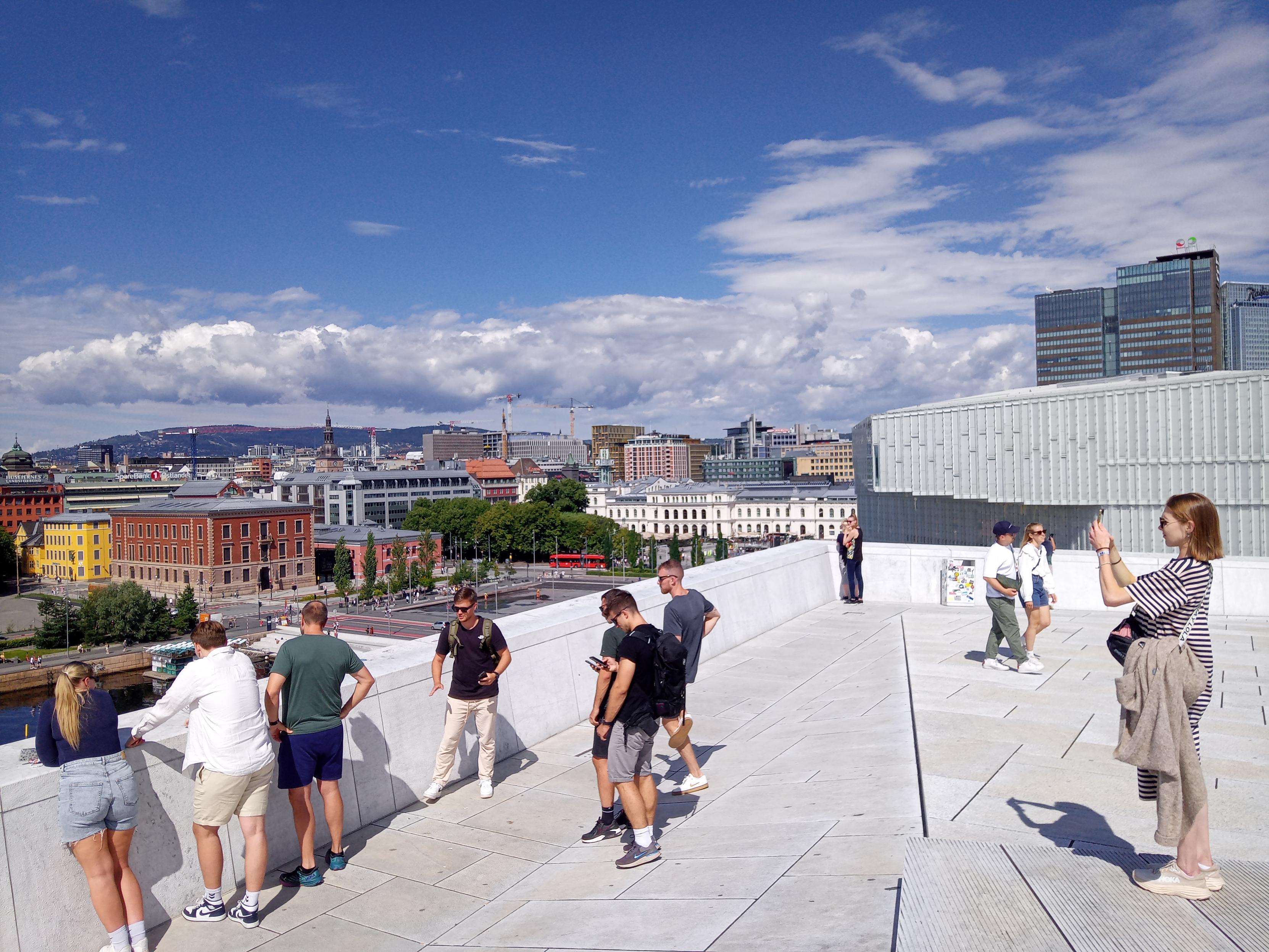 Photo taken from the roof of the opera house in Oslo and towards north. A light greyish white surface high up, people strolling around, looking at the view and taking photos. In the background the upper part of the library, also a white building. In the far back the white building that is Oslo Sentralstasjon, Oslo Central Station and other buildings north of the opera house. Blue sky with some white clouds particularly at the horizon. Everything in the picture is sunlit.