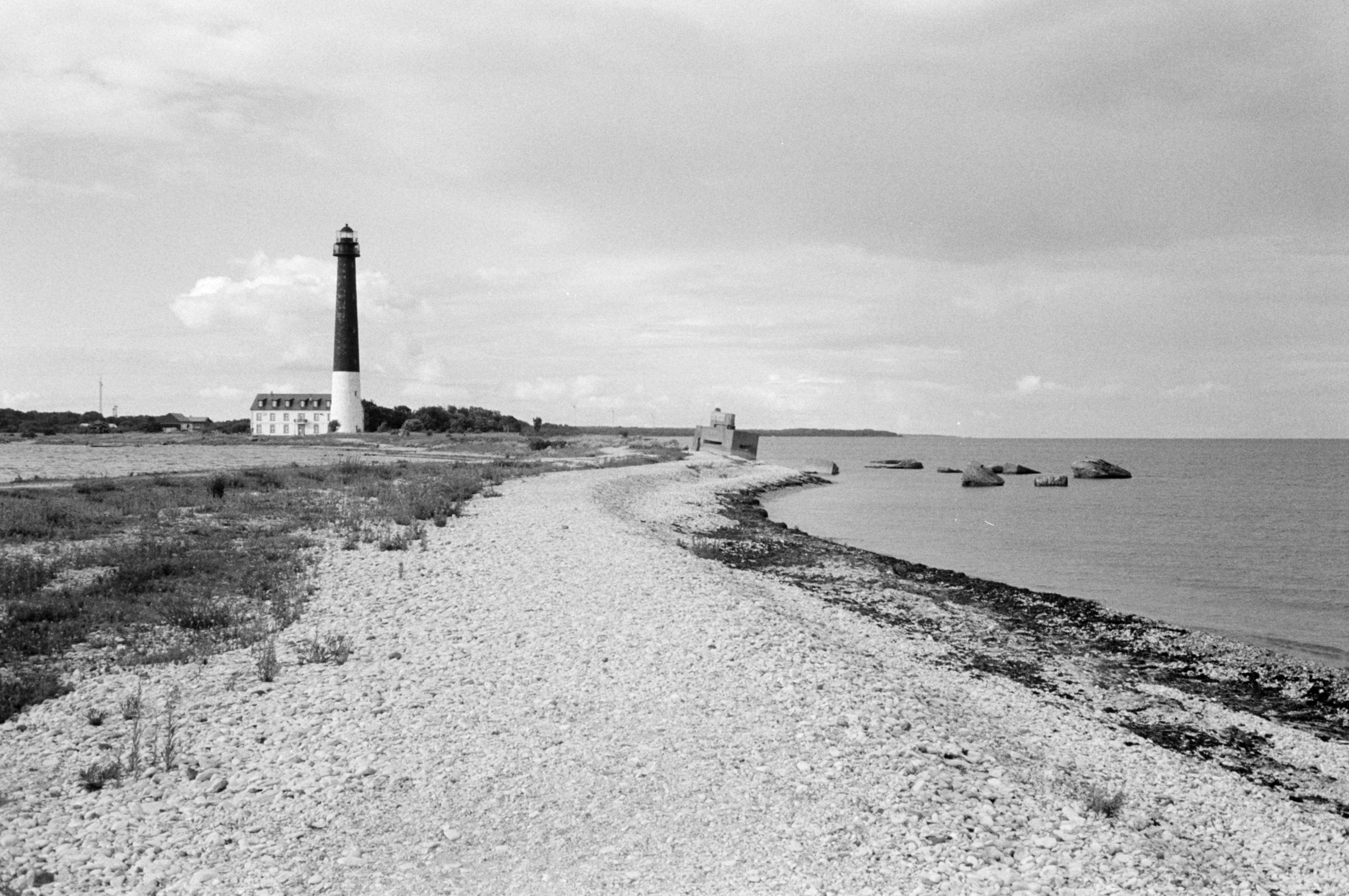 Schwarz-Weiß-Fotografie

In der Bildmitte ein steiniger Strand, der auf einen schiefstehenden Bunker im Hintergrund zuläuft. Rechts das Wasser. Links im Hintergrund ein Leuchtturm, dessen unteres drittel weiß gestrichen ist, die oberen zwei drittel schwarz. Am Leuchtturm schließt ein zweistöckiges weißes Haus an.