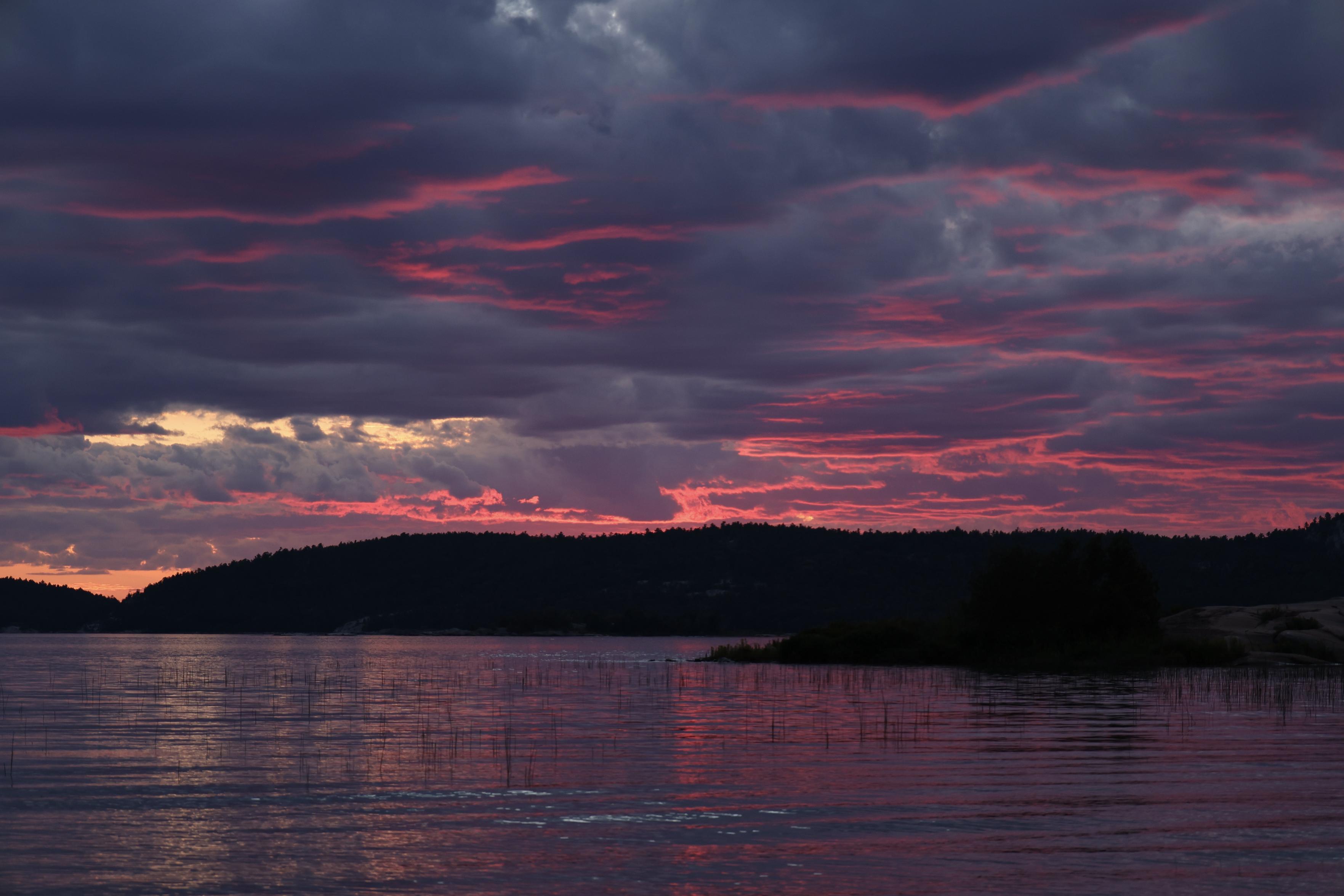This is a landscape format photo taken on a summer evening as a sunset over a lake was nearing its final stages. The clouds had darkened and traces of pink colouring were visible. The refection on the lake shimmered as gentle waves came ashore.