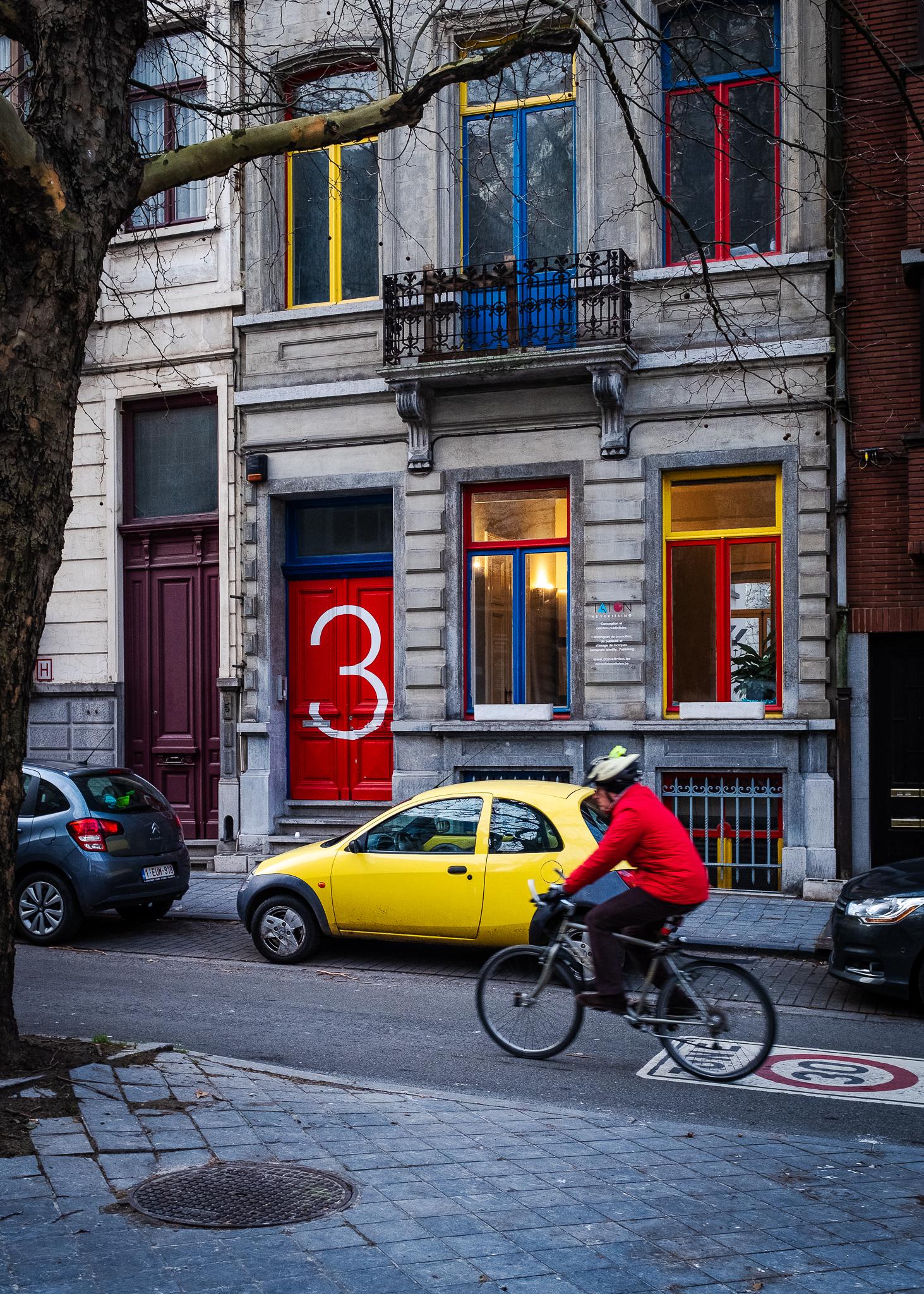 A cyclist with a red jacket is passing by a yellow car parked next to a red door with a very big number 3 painted in white on it a little street in Brussels.