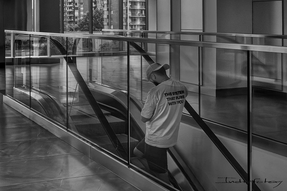 A Man Wearing a T-shirt saying: 'The System That Runs You" is going up in an escalator.