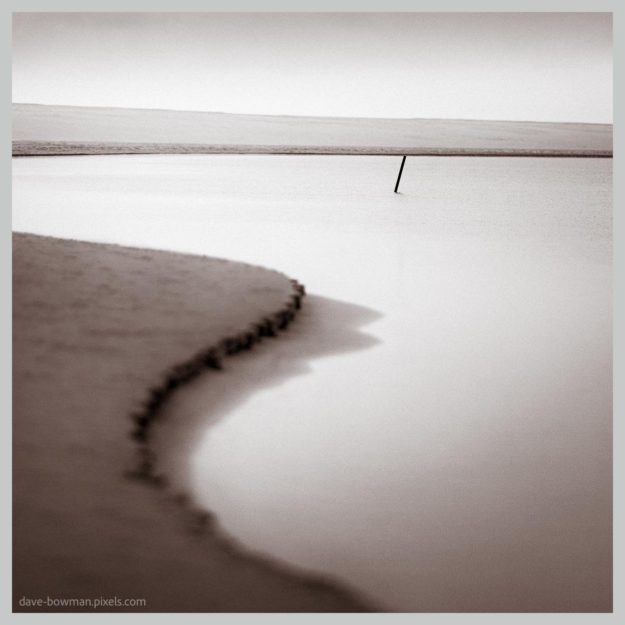A photograph of Kijkduin Beach in the Netherlands, shown in monochrome with a semi-abstract composition of a smooth, shallow pool of water stretching across the foreground and a single piece of wood protruding from its surface. Above it, an open sky occupies much of the image, with the simple forms and uncluttered composition creating a quiet, contemplative atmosphere.