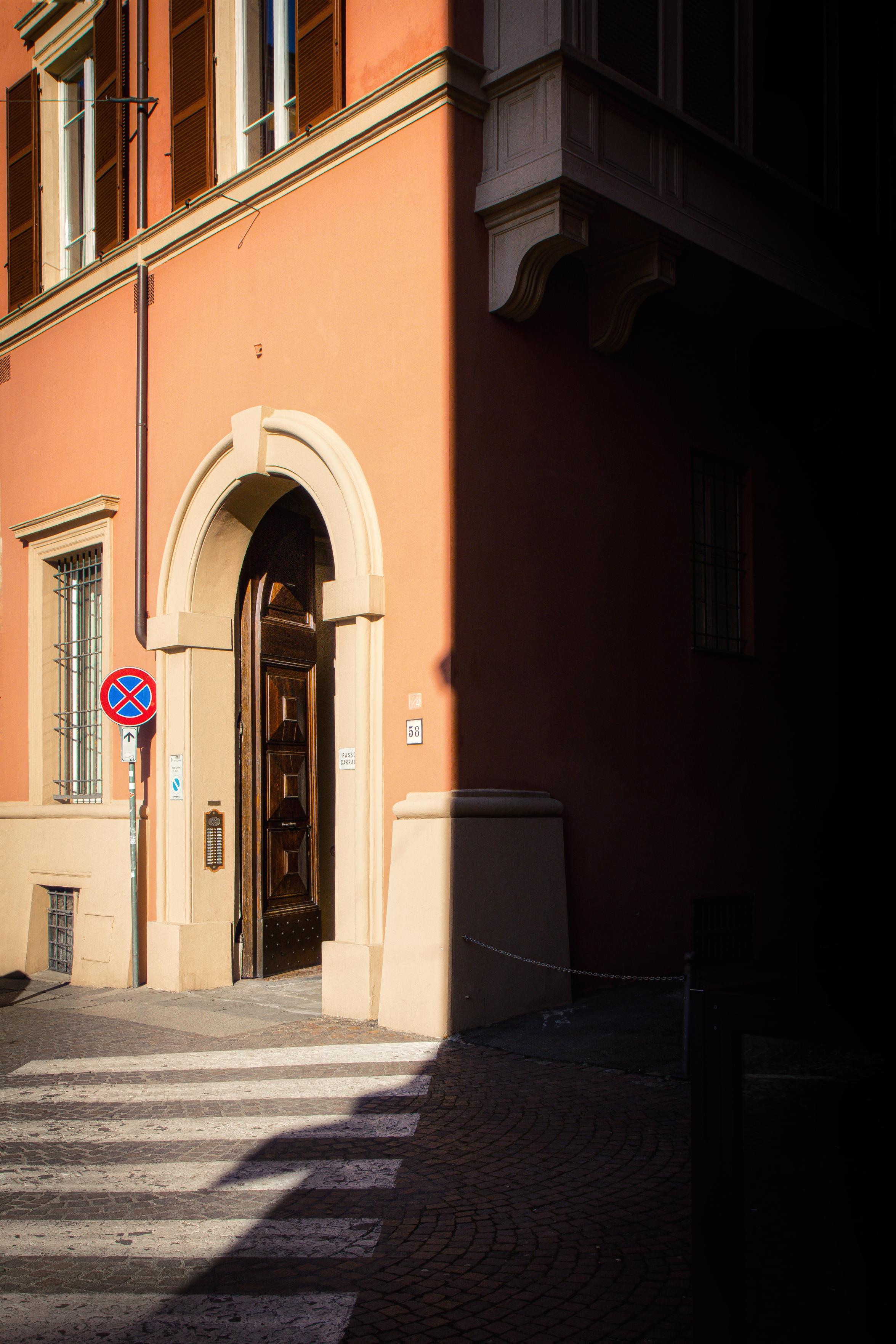 A photo of a door in via tagliapietre. It Is taken from the corner. The right hand side is covered in shadows while the left hand side is in birhft sunlight. There is an open, brown wooden door in a stone arch. Beside it is a road sign and coming up towards the door is a pedestrian crossing.
