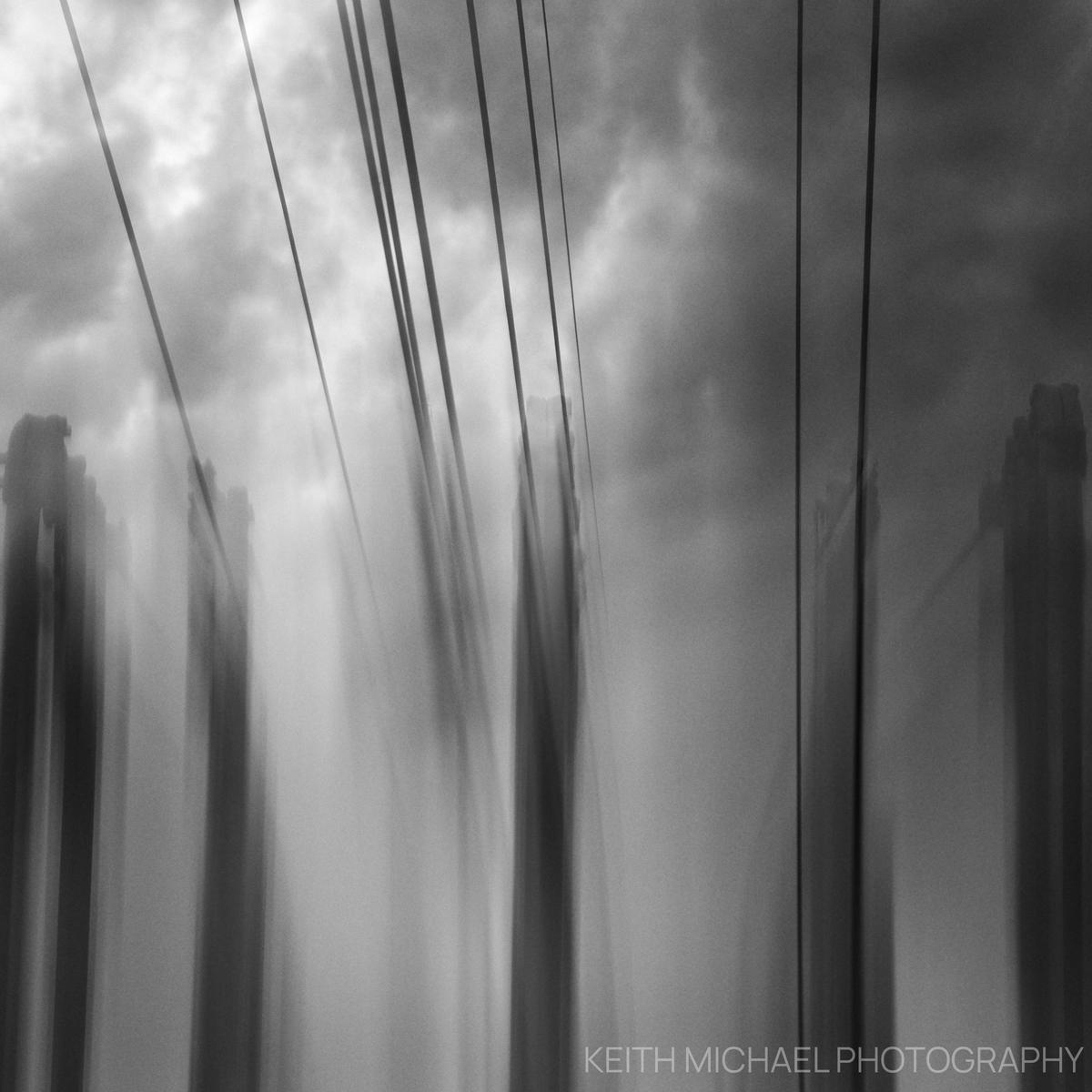 Double exposure black and white photograph with power lines converging on pillars at a substation. There is a moody cloud covering behind it.