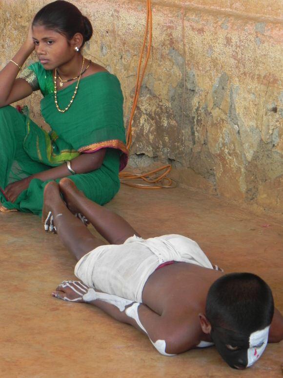 A woman in a green sari sits against a worn wall, looking away with one hand on her head, while a child lies face down on the floor in the foreground with black-and-white face paint and a white cloth around the waist.