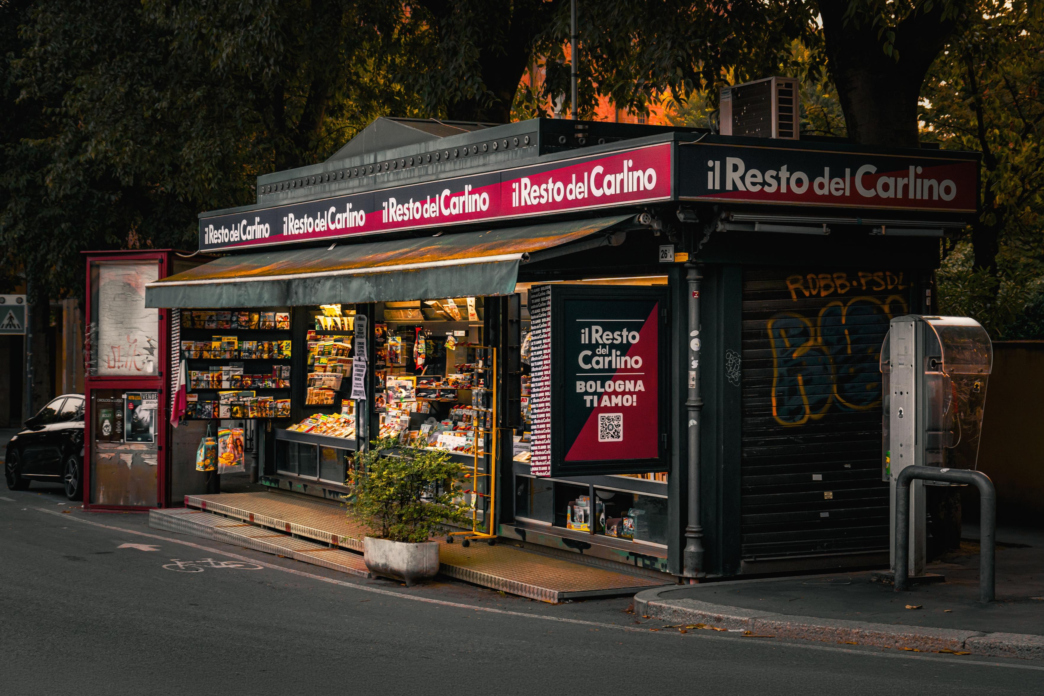 This is a photo of an outdoor newsstand. It has magazines, newspapers, and various small bits and pieces. It sits along a street under shaded trees. There is bright red branding for the Italian newspaper "il Resto del Carlino" which  lines the upper part and side panel of the kiosk. On the left there is a red poster stand and on the right there is a  grey payphone box with a metal guard.