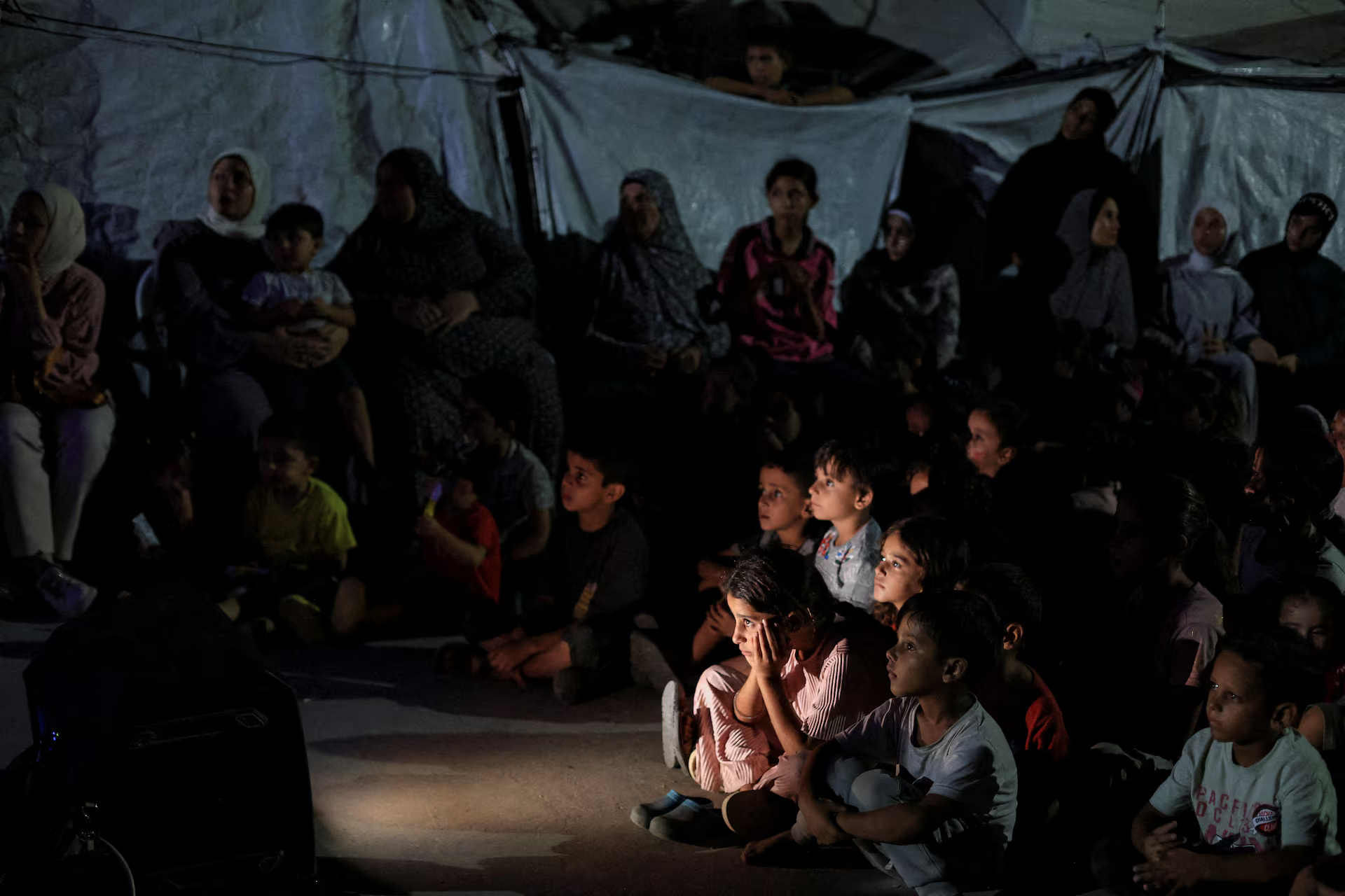 Palestinian children watch a cartoon film projected on a screen during a mobile cinema screening at a tent camp.