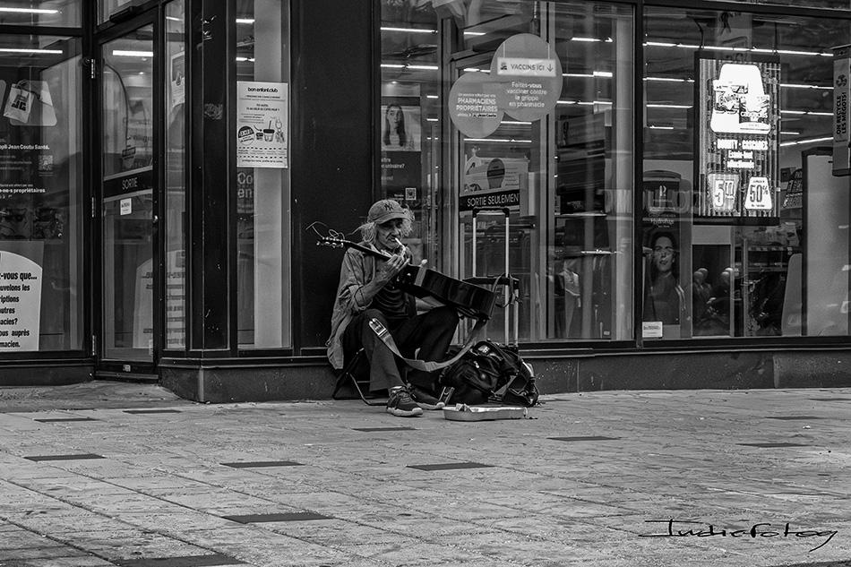A street musician, seemingly living in dire conditions, is looking at his guitar while sitting on the sidewalk.