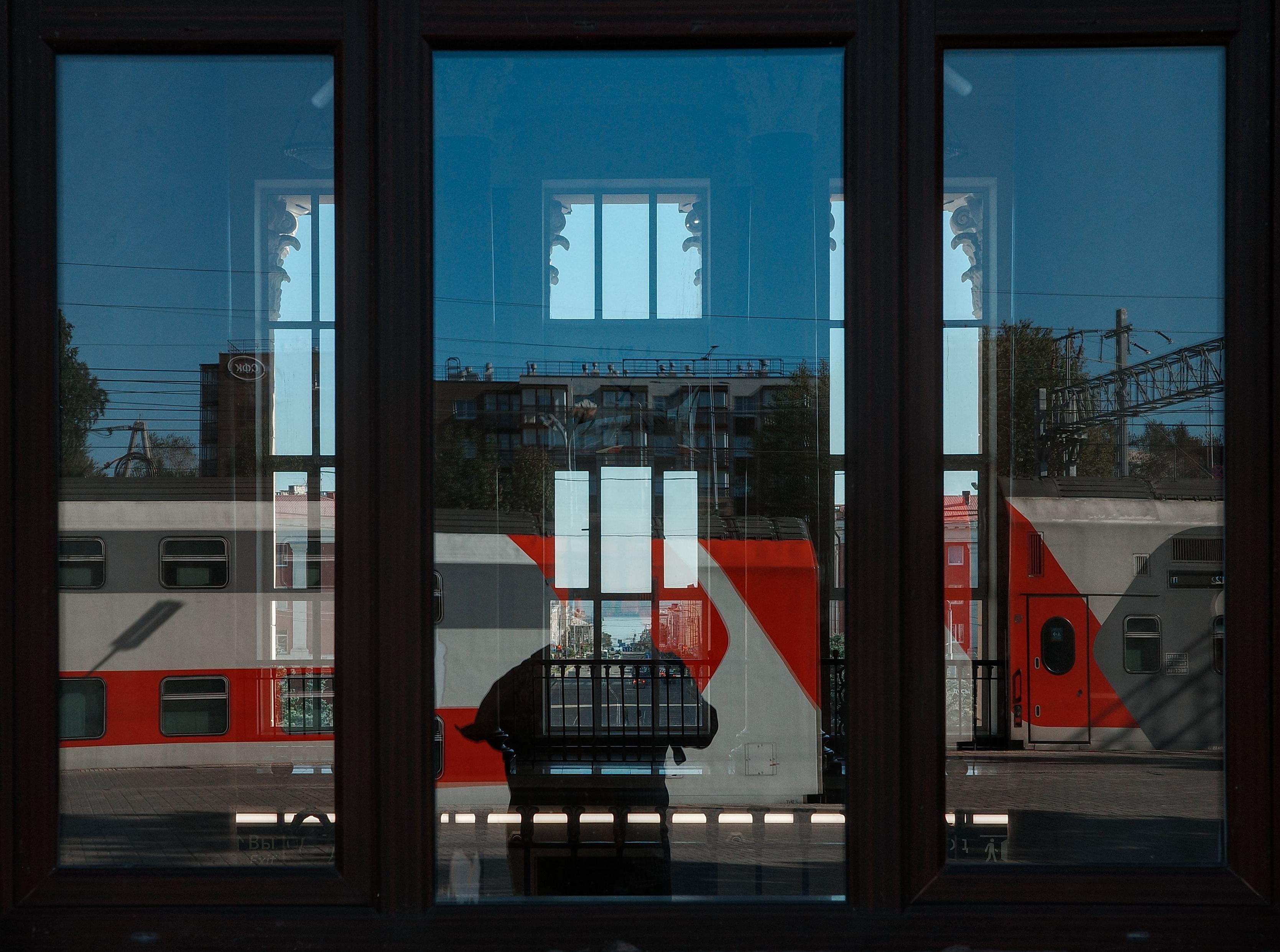 This color photograph captures a reflection in the window glass of a large pavilion. Three dark window frames create a strict rhythm, while the glass reflects a railway station: red and gray train cars are visible outside, along with fragments of neighboring buildings. In the central window, the facade of a house with white columns is reflected, with a clear blue sky visible behind it. In the lower part of the reflection, like a shadow, the silhouette of a person or object appears, adding graphic quality and depth to the frame. The image conveys a sense of a frozen moment, where architecture and transport create a complex, layered composition.